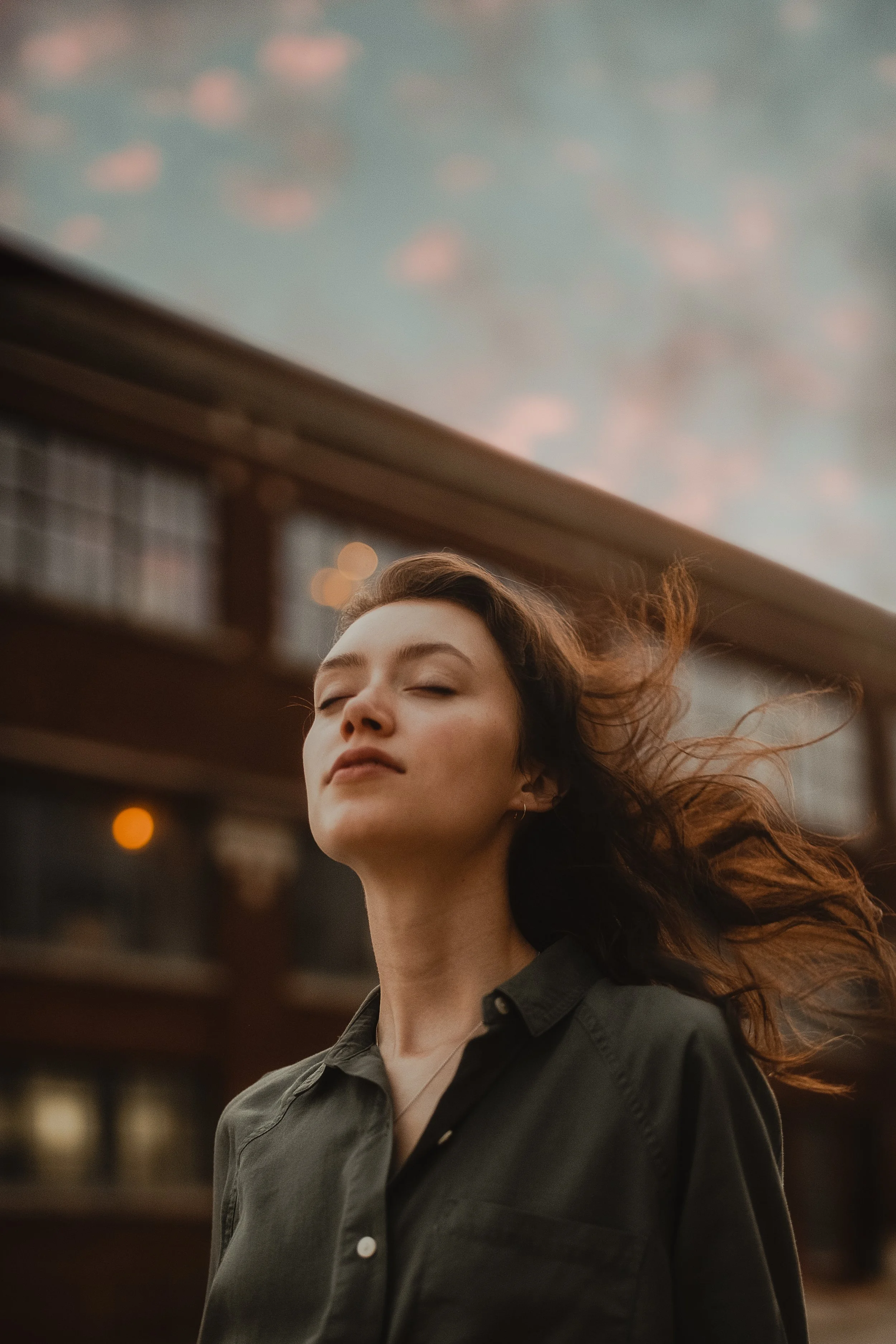 Young woman with closed eyes outdoors, wind blowing her brown hair, wearing a dark green button-up shirt, against a blurred urban background.