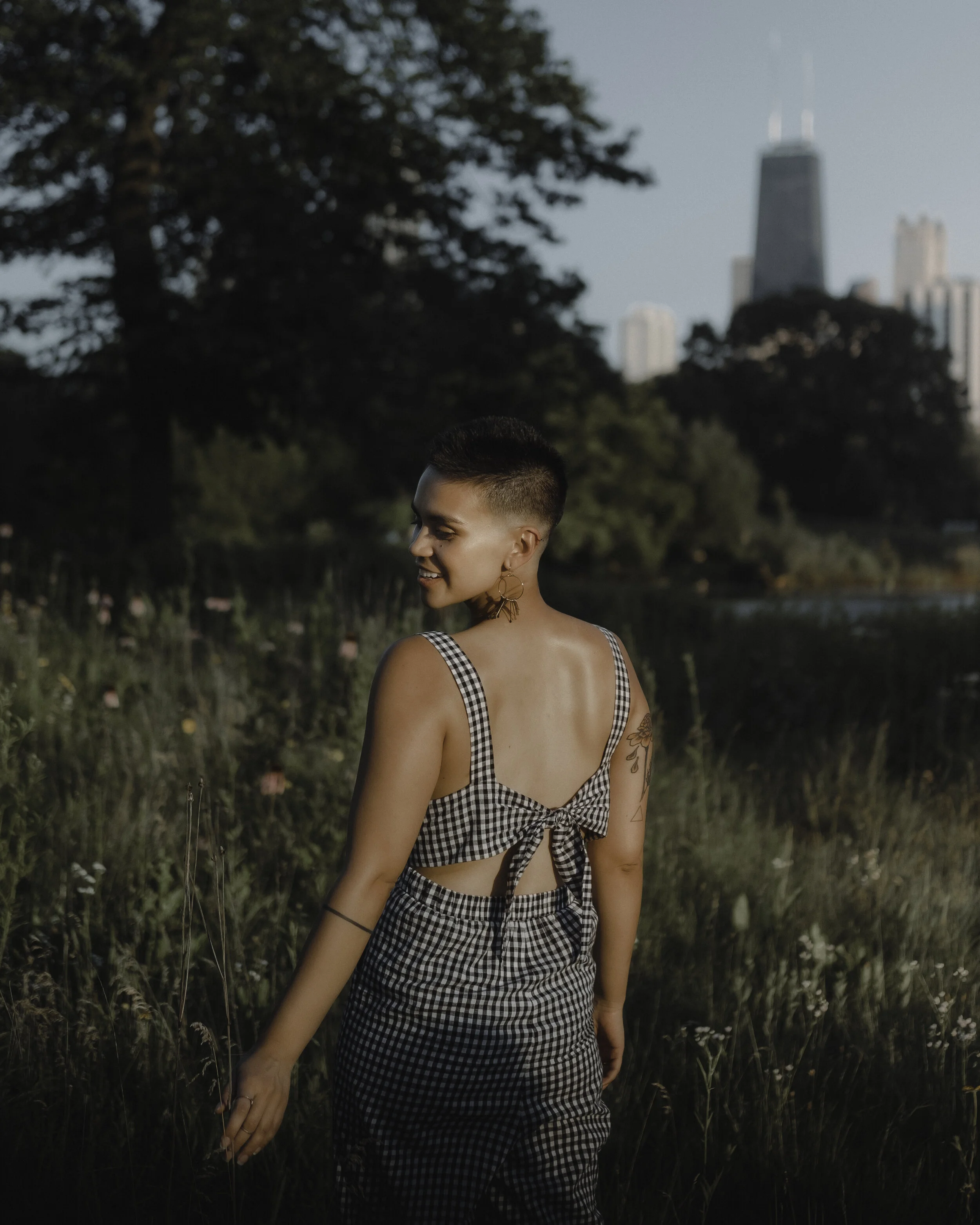 A woman with short hair and tattoos on her left arm, wearing a black and white checkered crop top with a tie in the back, and matching checkered pants, walking through a grassy area near a lake or river, with trees and a city skyline with tall buildi