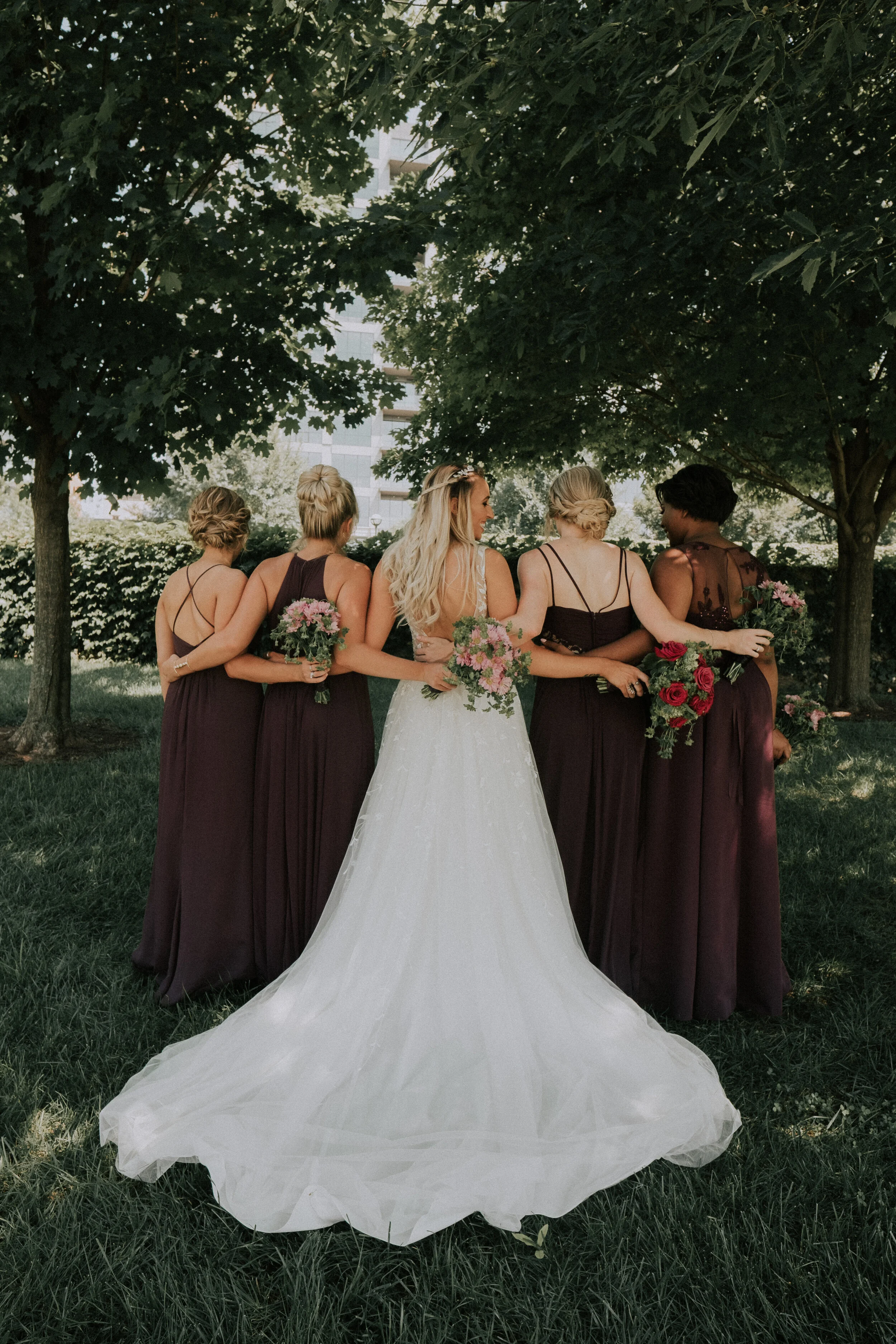 Bride in a white wedding dress with blonde hair and four bridesmaids in dark purple dresses, holding bouquets of flowers, standing outdoors under a large tree.