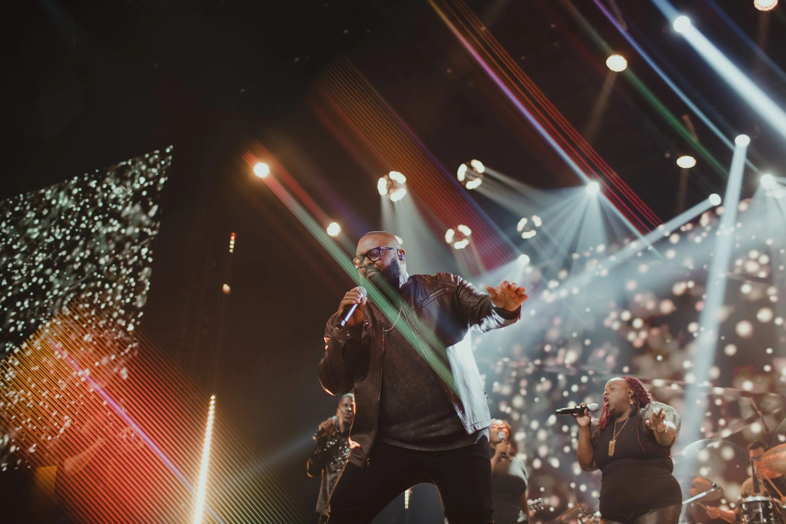 A man with glasses and a beard, wearing a leather jacket, singing into a microphone on a stage with colorful lights and digital screens, accompanied by backup singers and band members.