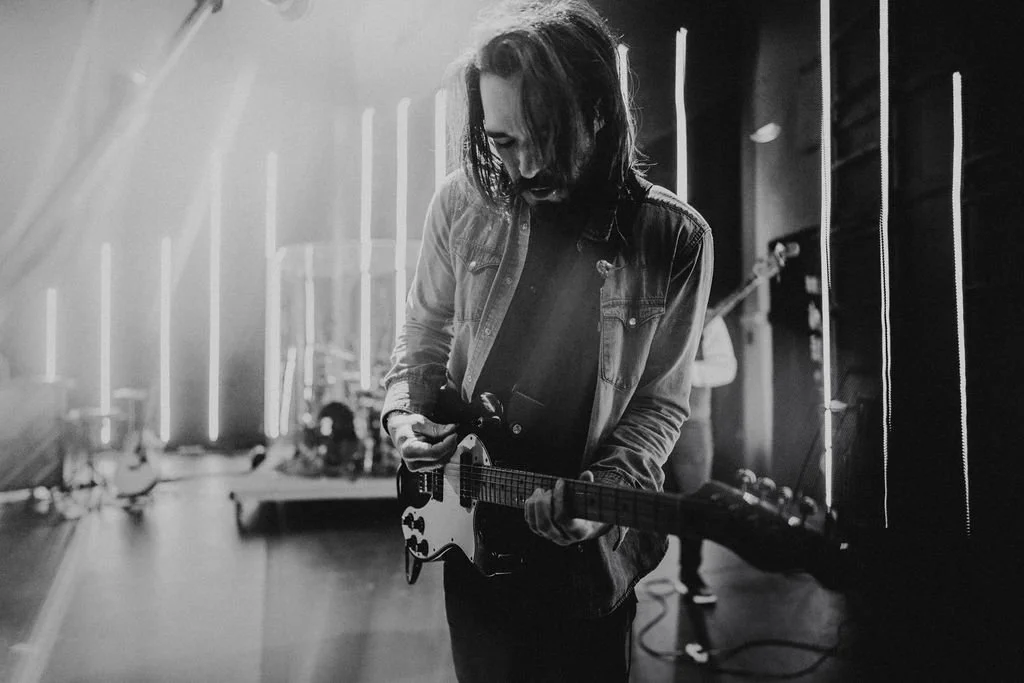 A man with long hair and a beard playing an electric guitar on a stage with vertical light strips in the background in black and white.