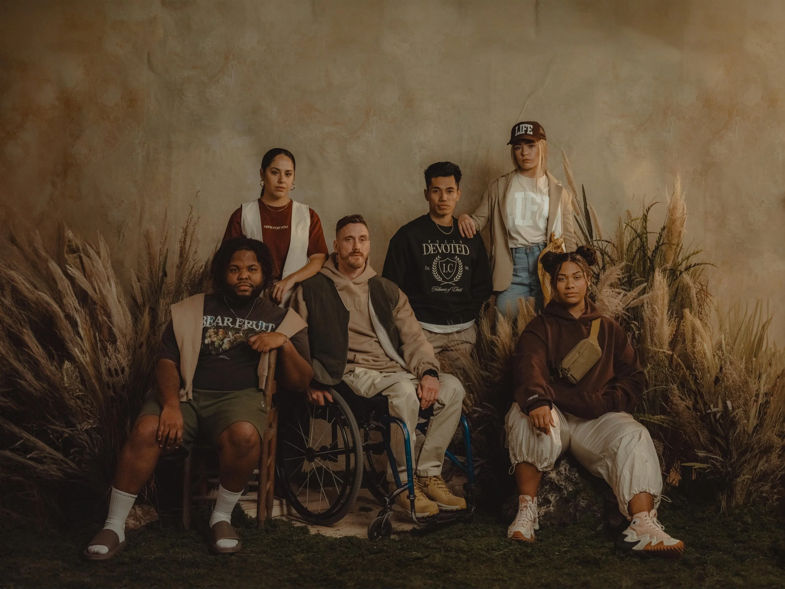 Group of seven diverse young people posing in an indoor setting with tall grasses and a beige background.