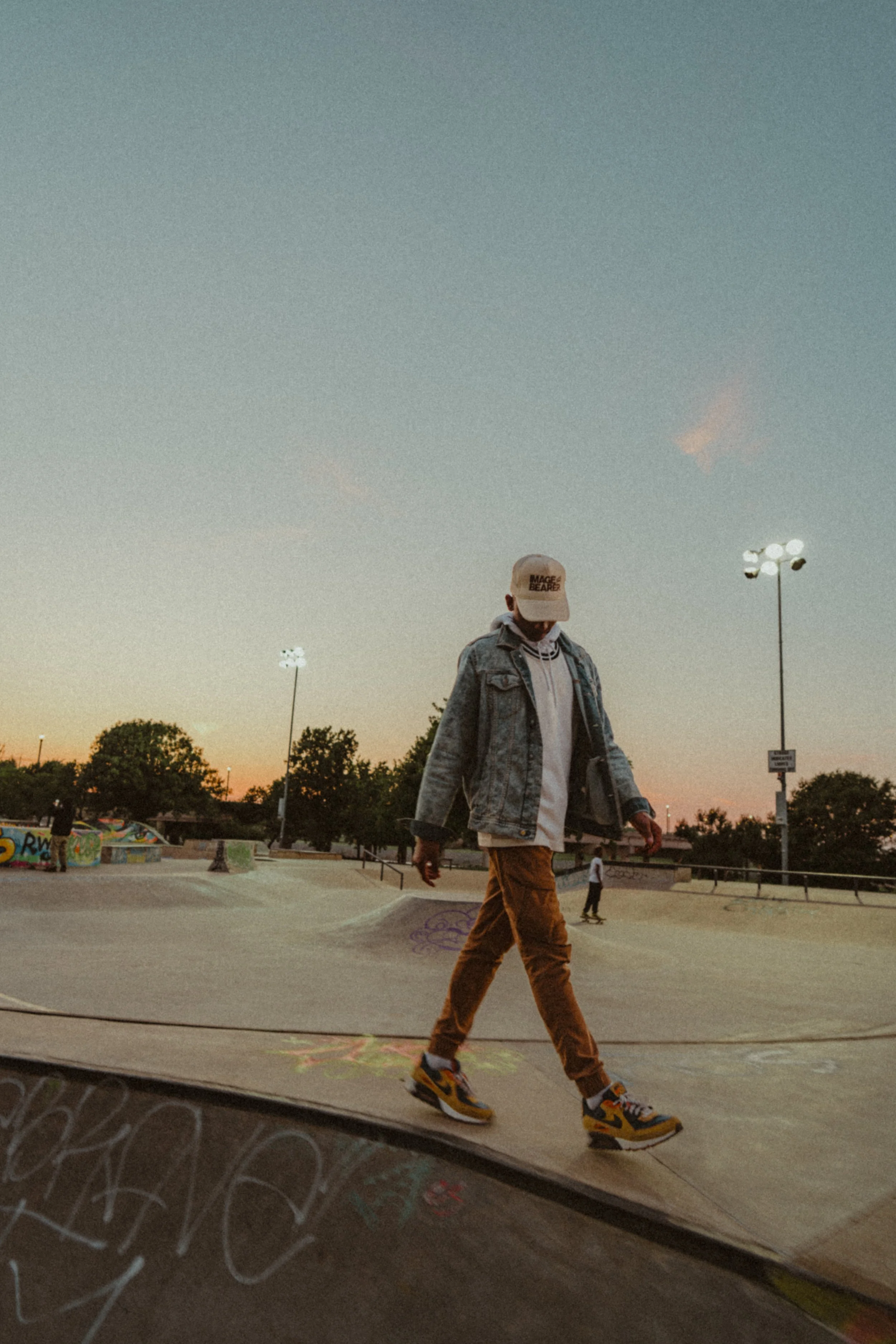 A person walking at a skate park during sunset, wearing a cap, denim jacket, hoodie, brown pants, and sneakers.