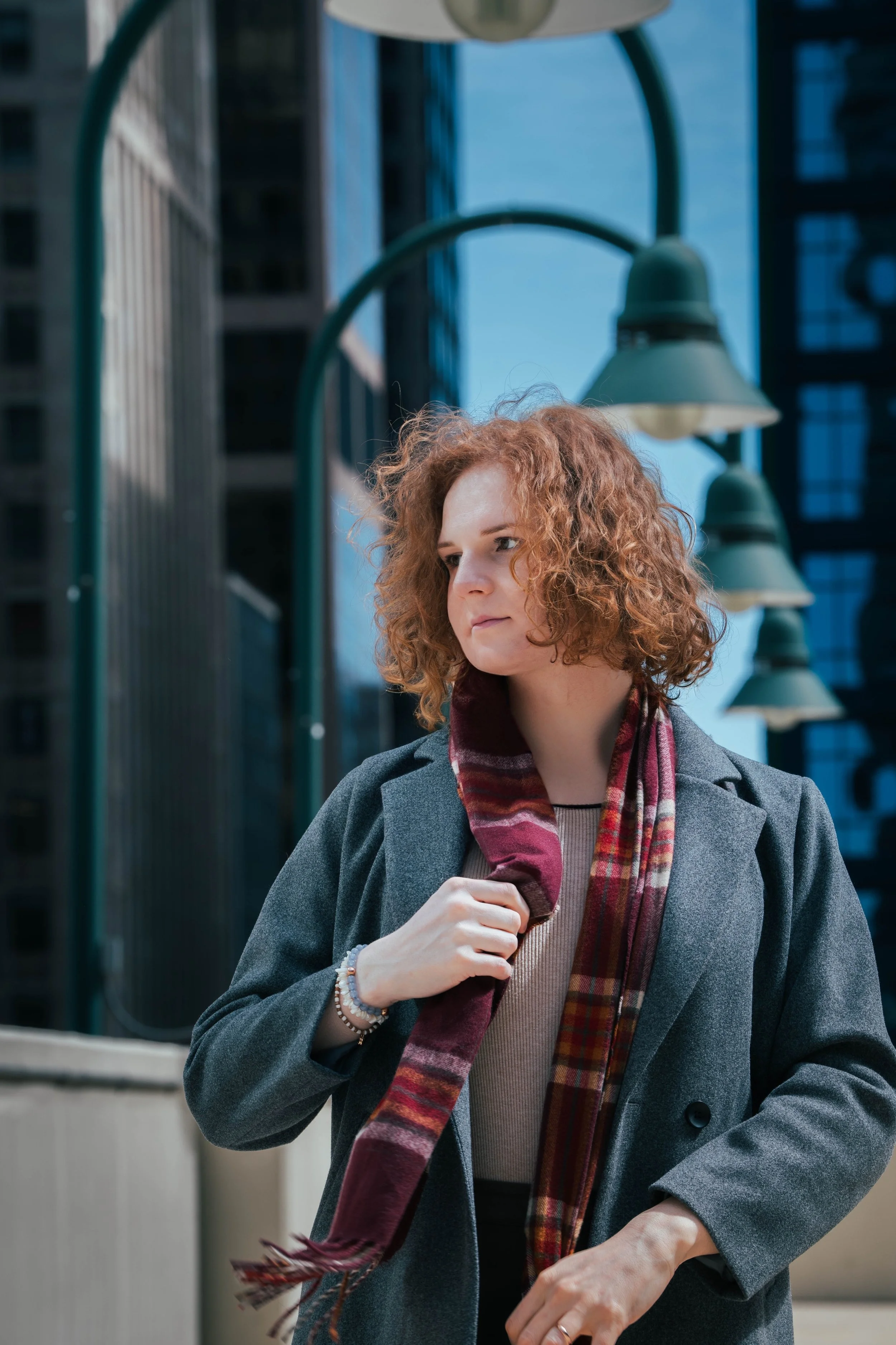 A woman with curly red hair wearing a gray coat and a plaid scarf outside in an urban area during daytime.