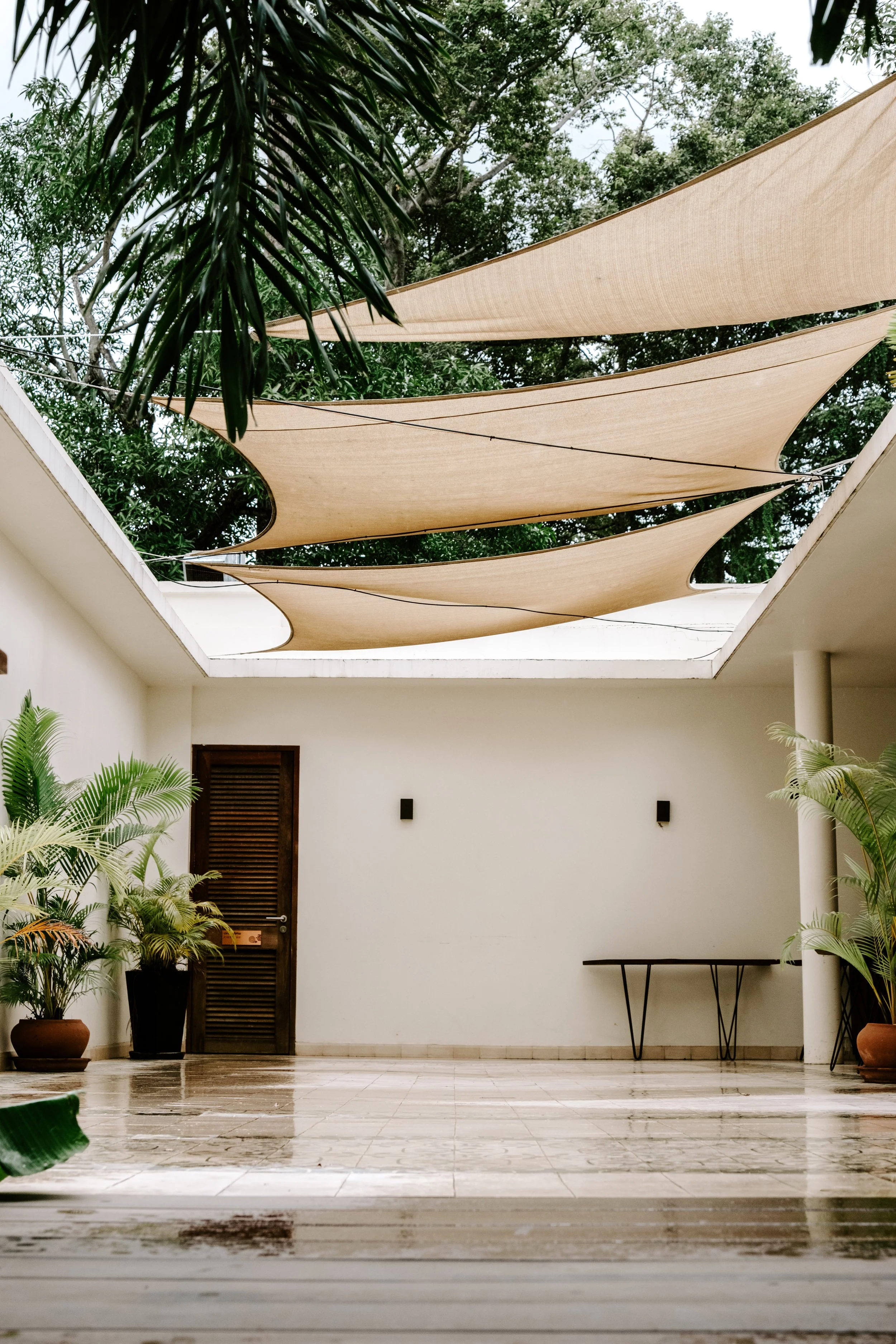 An outdoor patio with potted plants, a white wall with two black wall lights, a small black table, and fabric shade sails overhead.