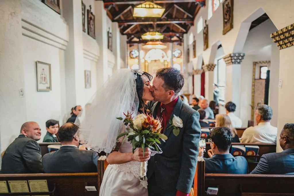 A bride and groom kissing inside a church, with guests seated in the background and the bride holding a bouquet of flowers.