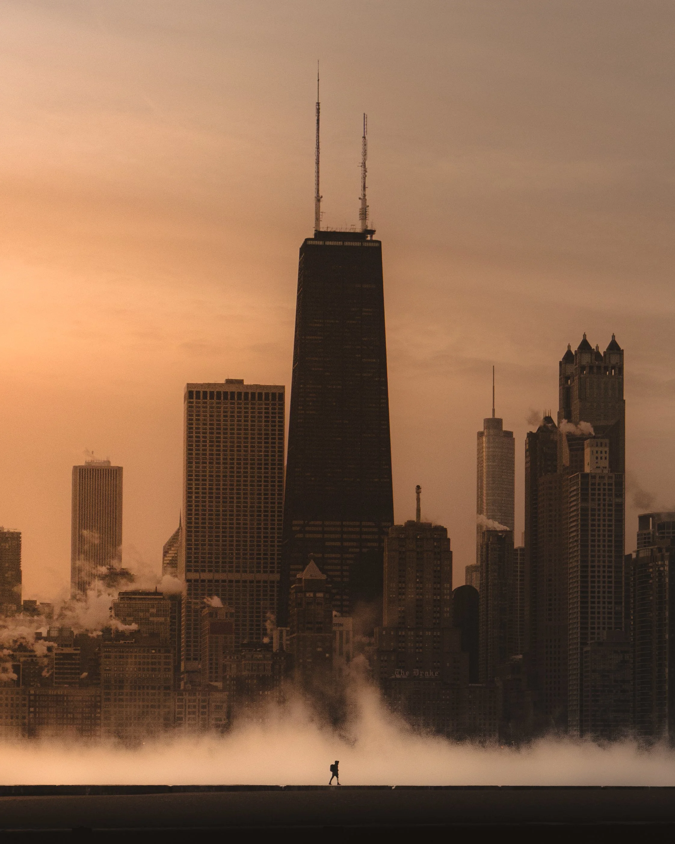 A person walking with a backpack in front of a misty city skyline at sunset, featuring tall skyscrapers including the tallest building with two antennas.
