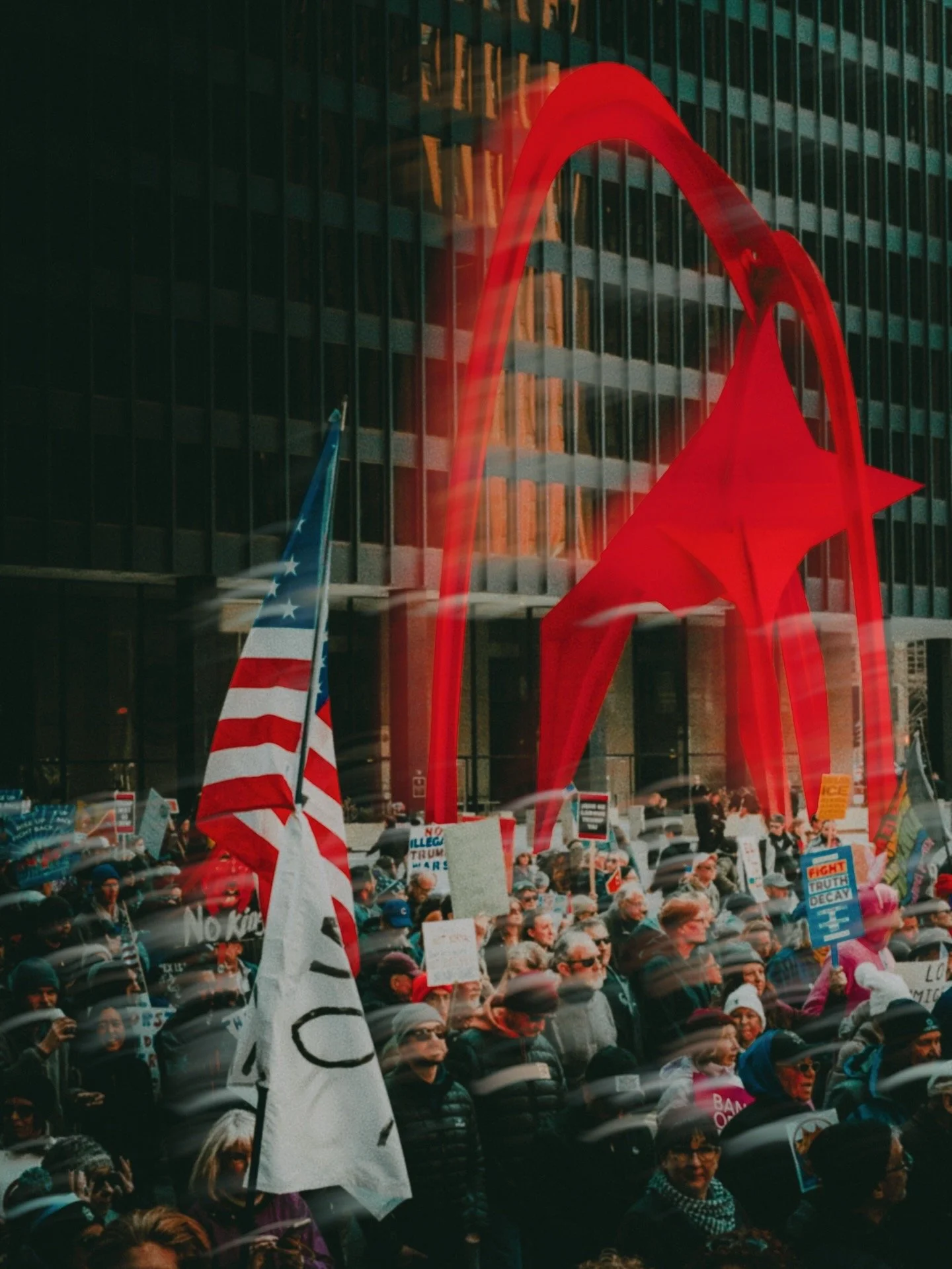 Crowd gathered outside a building with a large glowing red 'A' or arch symbol, holding signs and flags during a protest or rally.