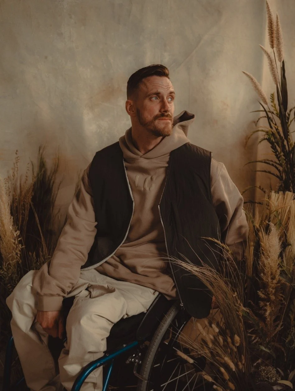 A man with a beard and short hair sitting in a wheelchair in front of a beige textured wall, surrounded by tall ornamental grasses and plants, wearing a tan hoodie and a black vest.
