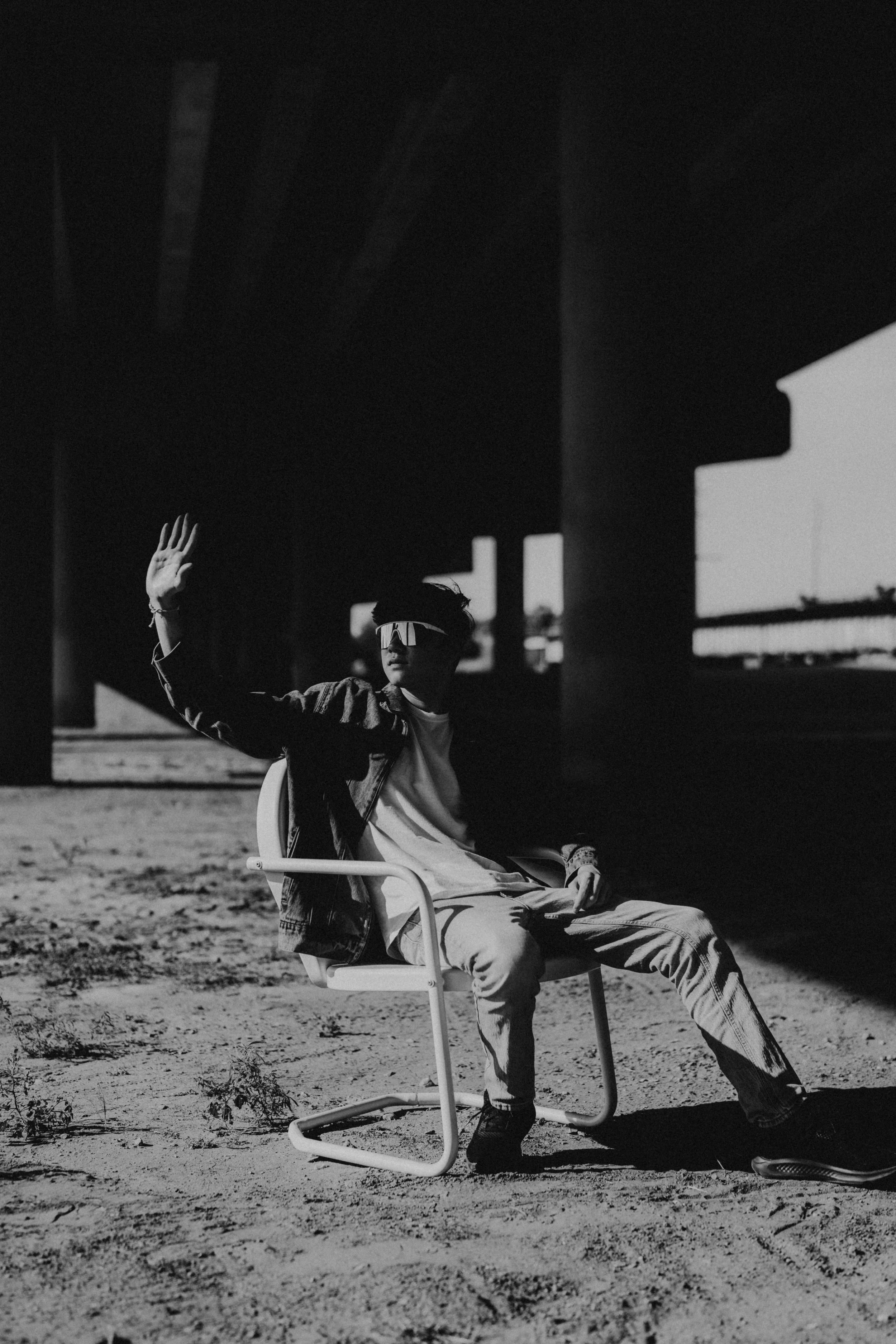 A person sitting on a chair outdoors in the shade, wearing sunglasses, a jacket, and casual pants, raising one hand in a waving gesture.