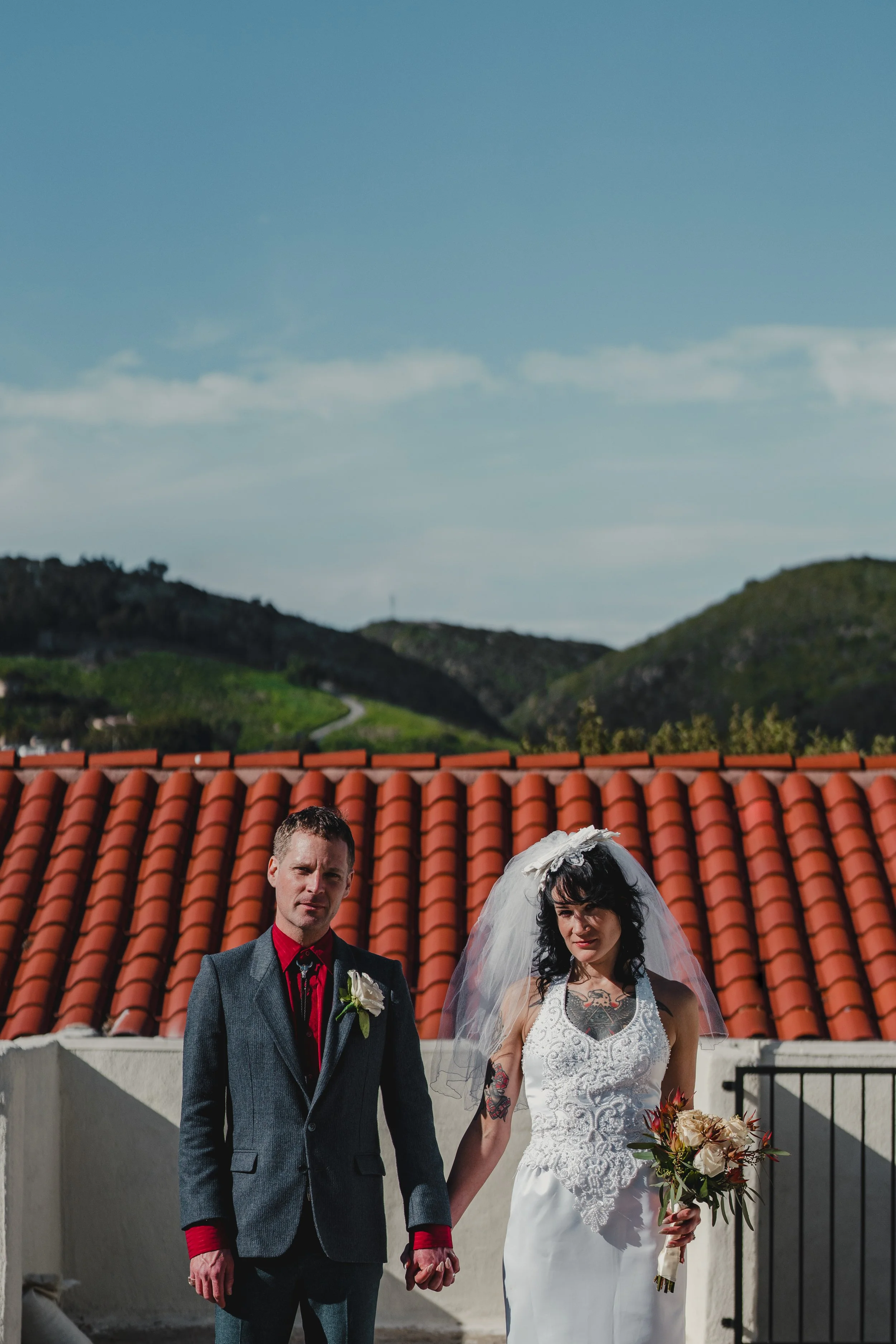 A couple standing outside, holding hands, dressed in wedding attire with a scenic backdrop of hills and a red-tiled roof.