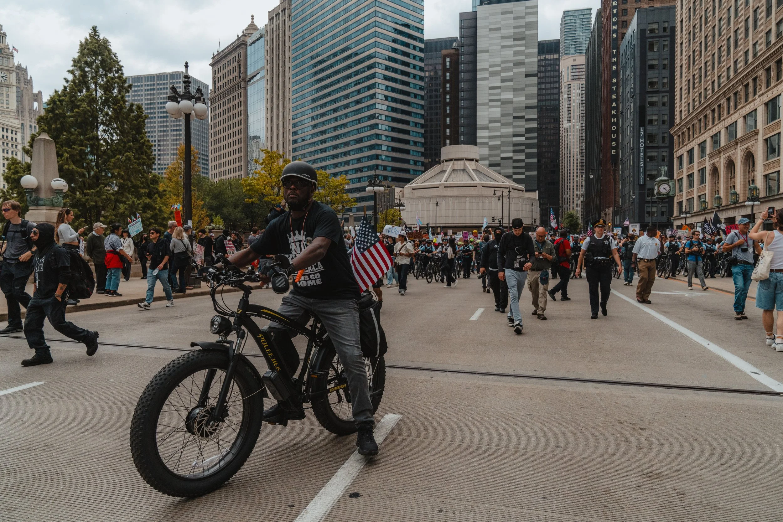 A man riding an electric bicycle in the foreground at a protest or demonstration in an urban city with tall buildings and a large crowd of people holding signs in the background.