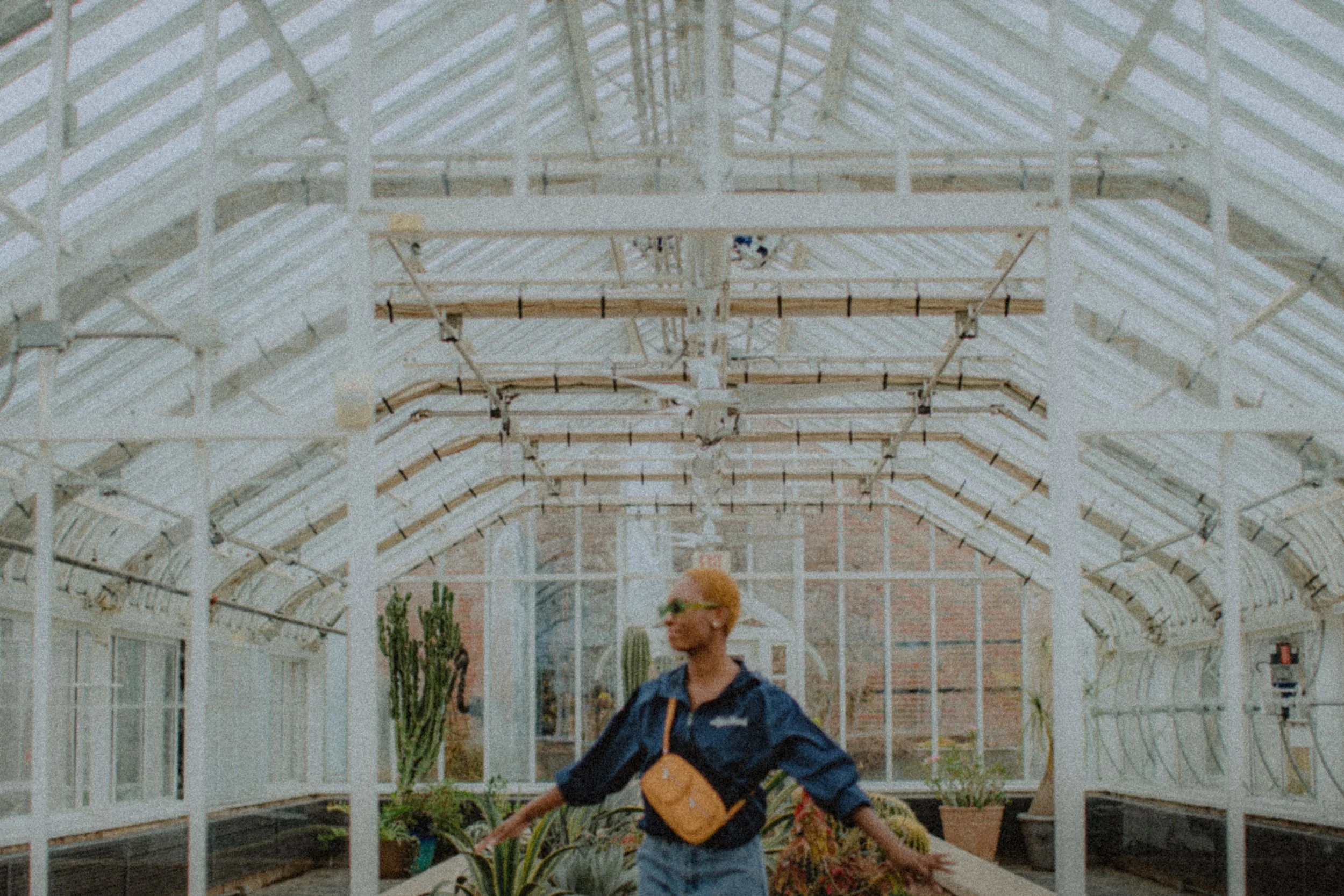 A woman with short blonde hair, green sunglasses, and a navy jacket with a tan crossbody bag, standing inside a greenhouse with plants and cacti.