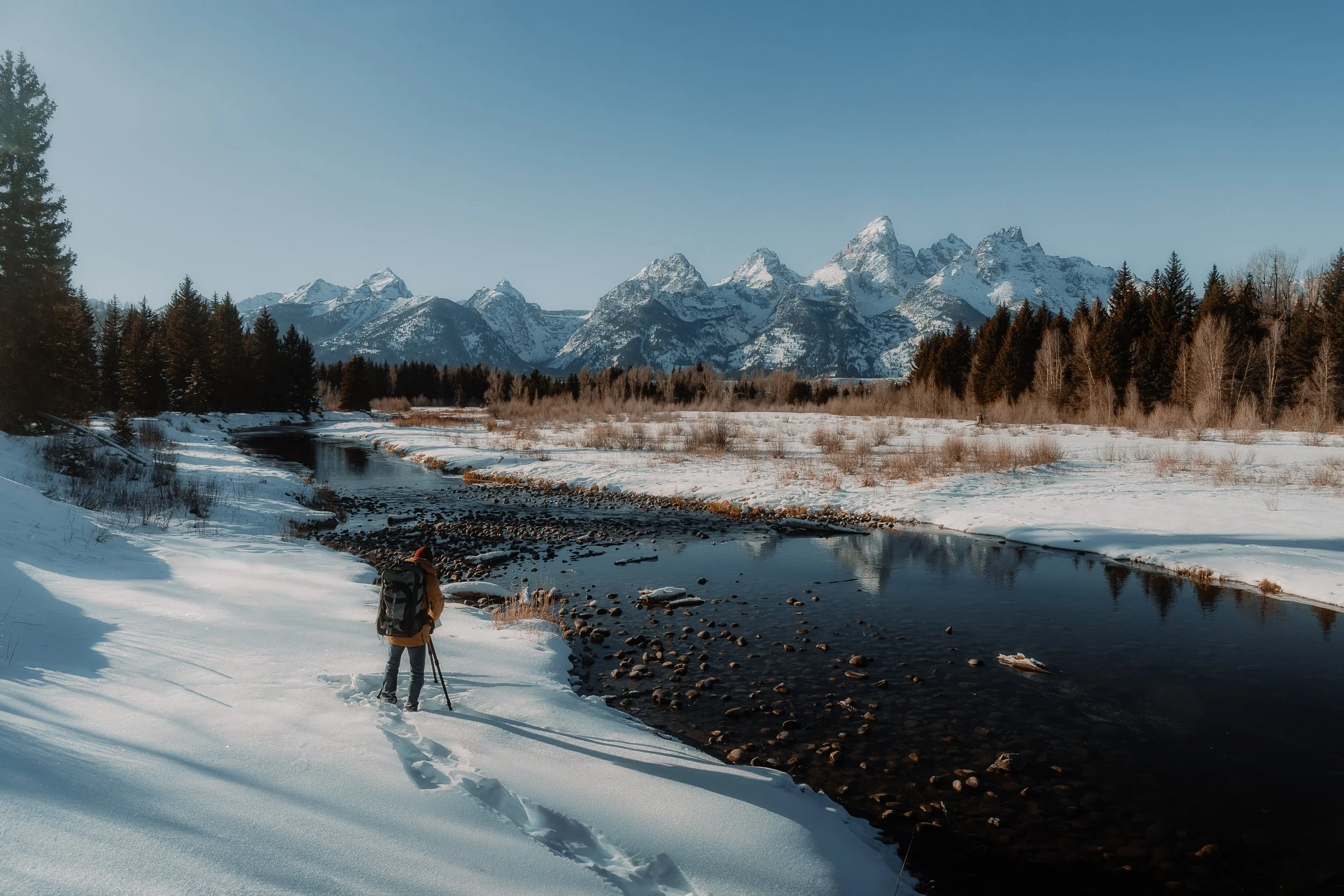 A person with a backpack and tripod standing on snow-covered terrain near a river, with snow-capped mountains and pine trees under a clear blue sky in the background.