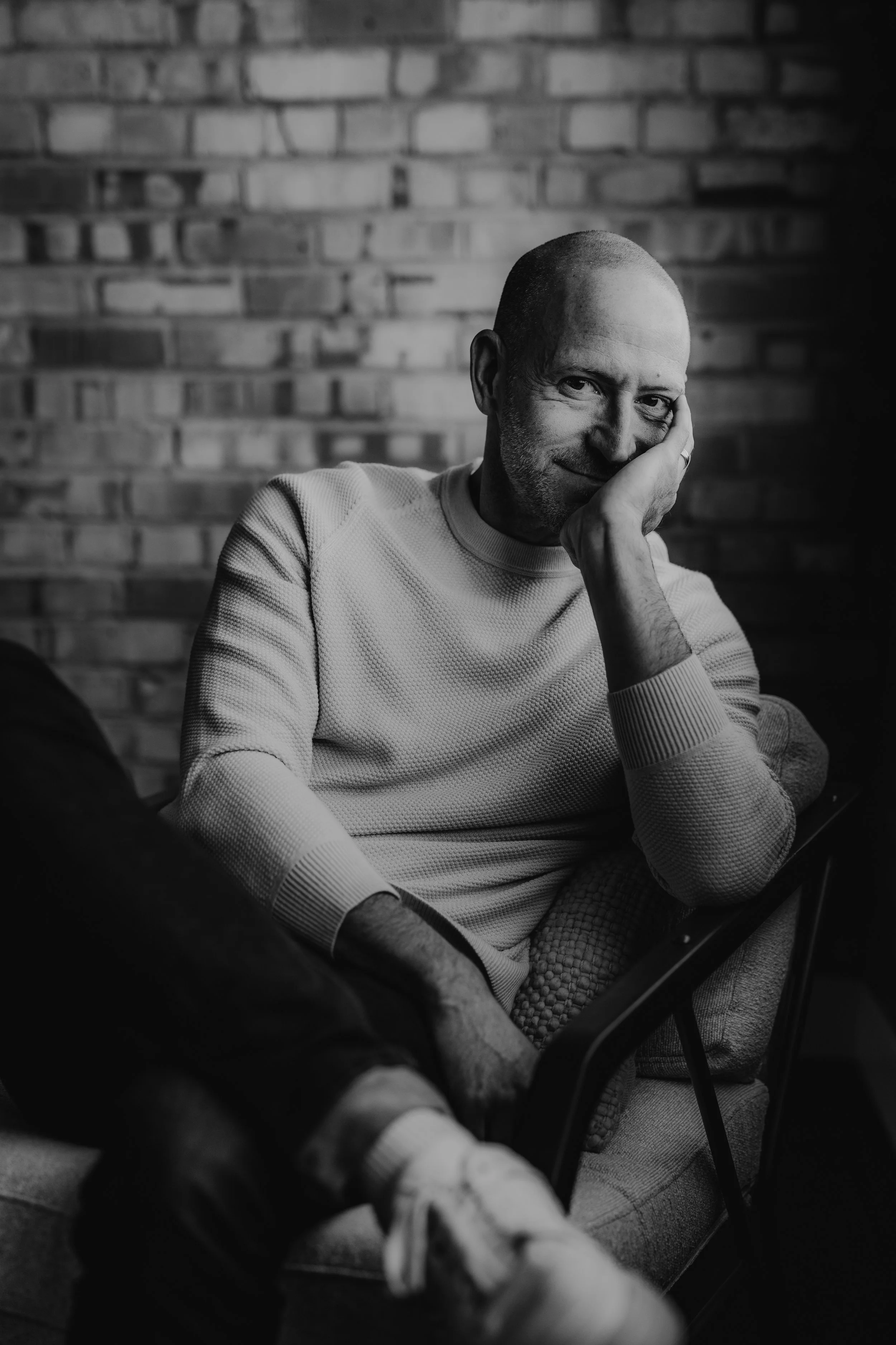 A black-and-white photo of a man with a shaved head, sitting on a chair against a brick wall, resting his head on his hand and looking at the camera with a slight smile.