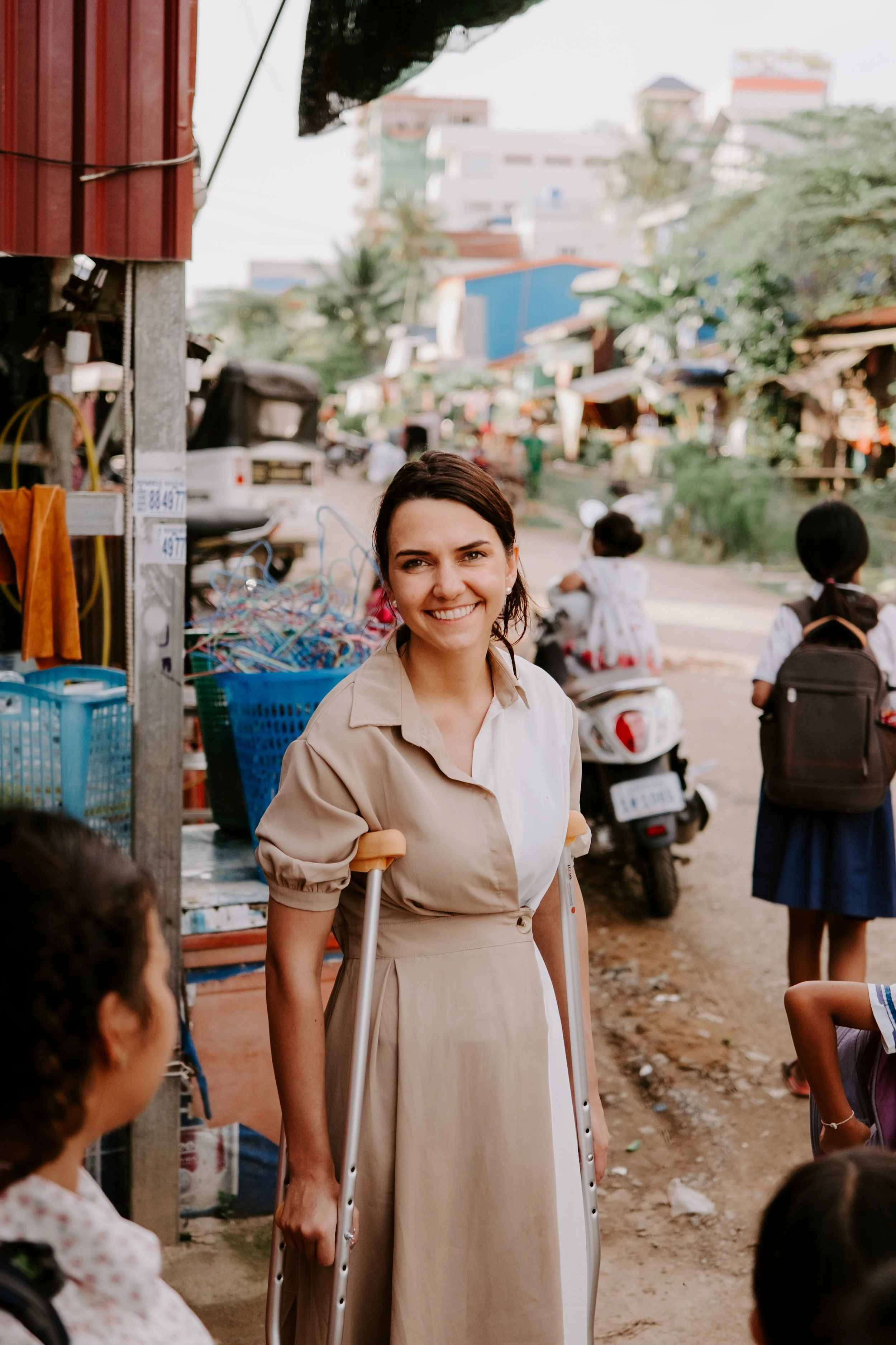 A woman smiling, using crutches, standing outdoors in a marketplace, with children and scooters in the background.
