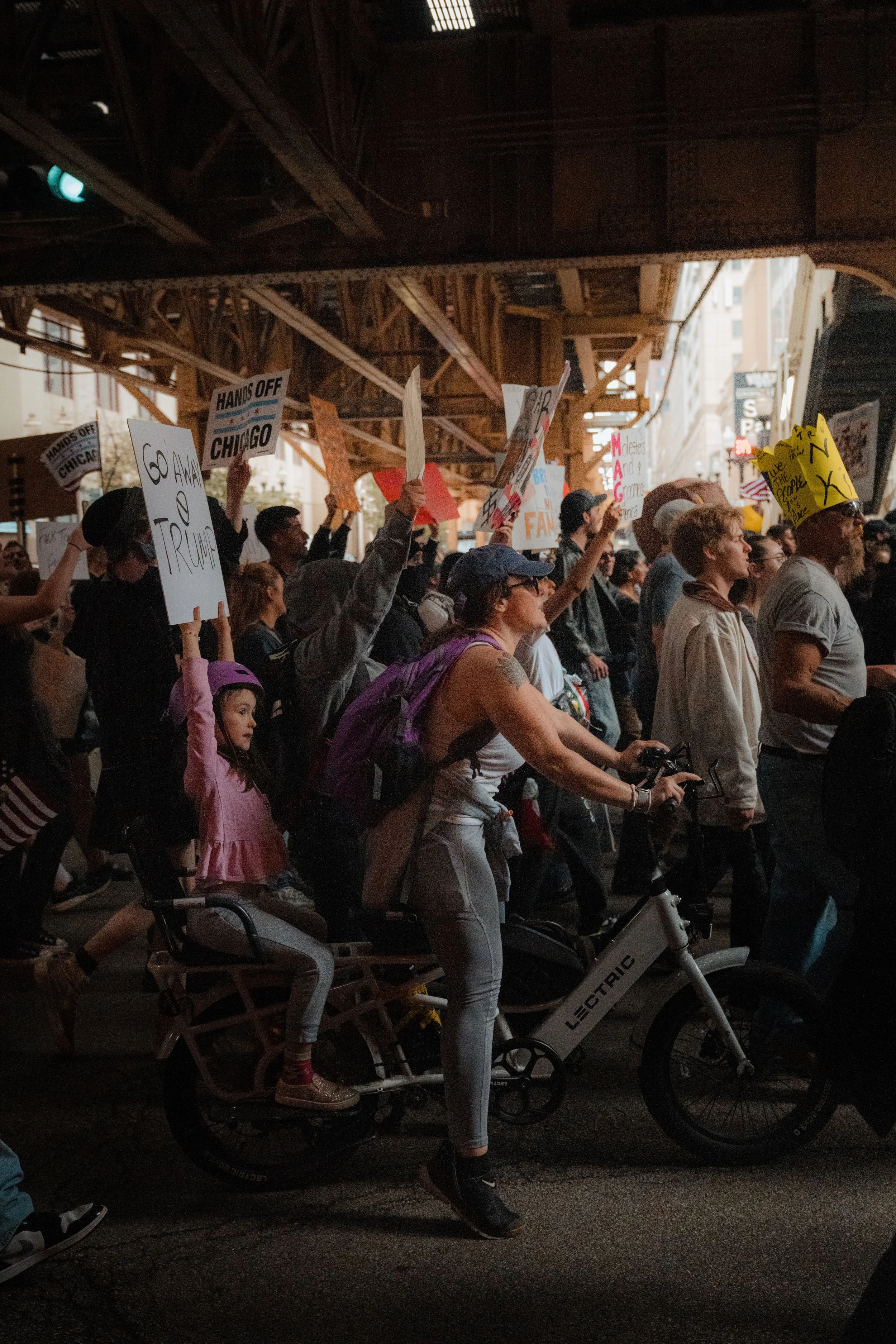 A crowd of protesters holding signs at a demonstration, with some signs mentioning Chicago, Trump, and other political messages, under an overhead structure in an urban setting.