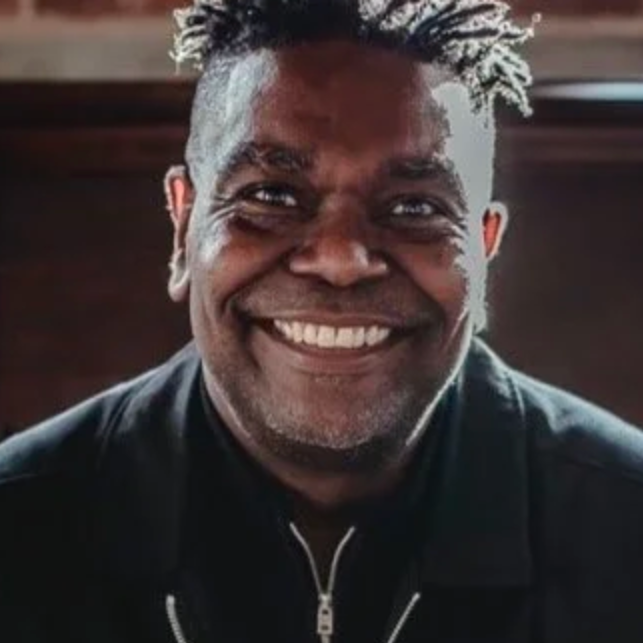 Close-up of a smiling man with short, spiked hair and dark clothing, indoors.