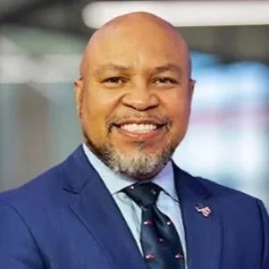 A smiling man with a beard wearing a blue suit, dress shirt, and tie.