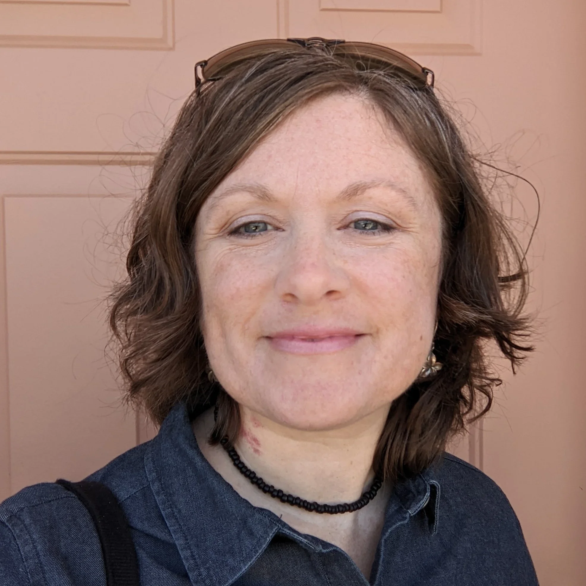 A woman with curly brown hair and light skin smiling at the camera, wearing sunglasses on her head, a black choker necklace, and a dark blue button-up shirt, standing in front of a pinkish background.