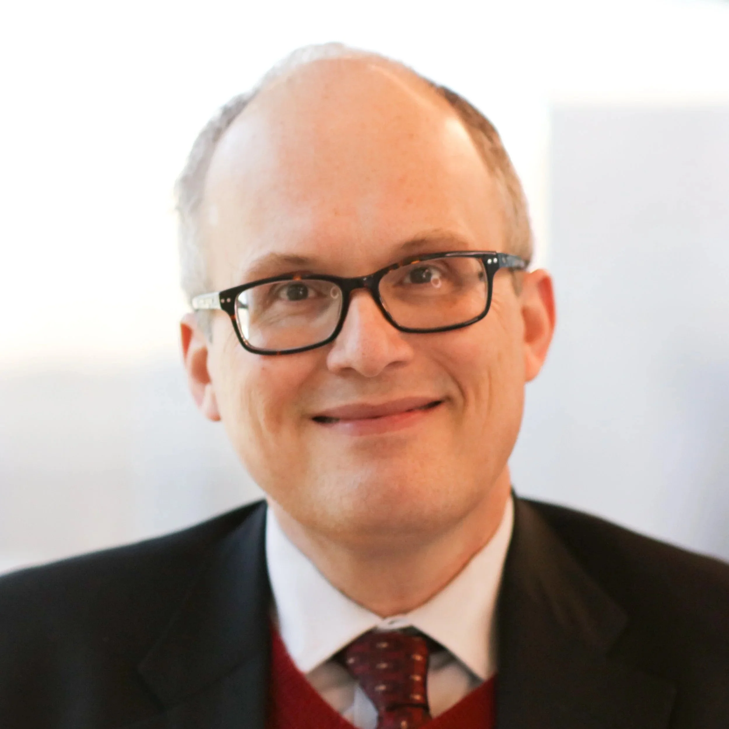 Portrait of a smiling man with glasses, wearing a suit, white shirt, and red tie.
