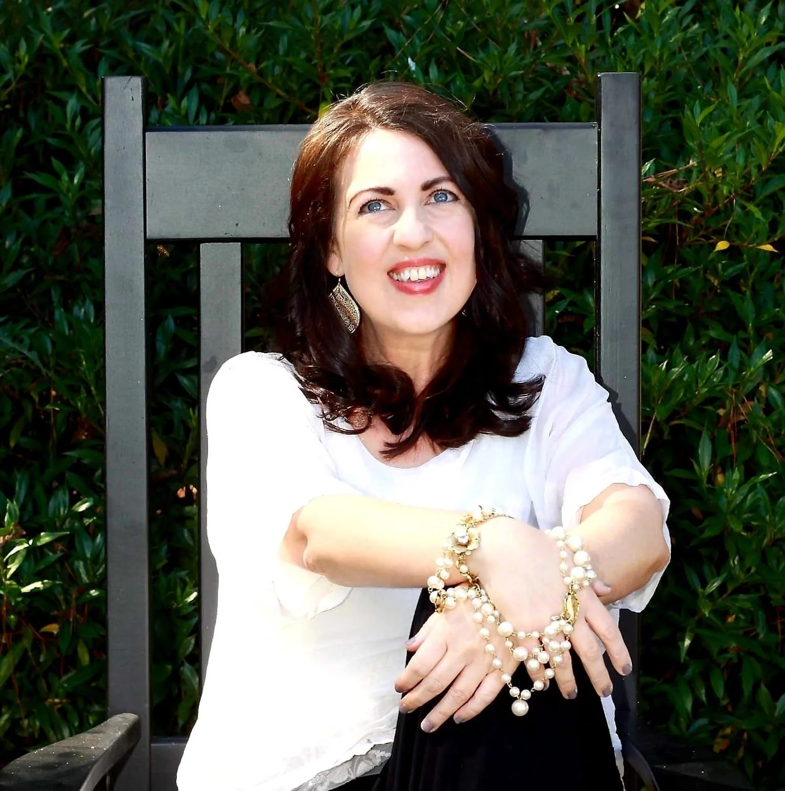 A woman with dark brown hair and blue eyes sitting on a black wooden chair outside, wearing a white top and multiple pearl jewelry pieces, smiling with arms crossed.