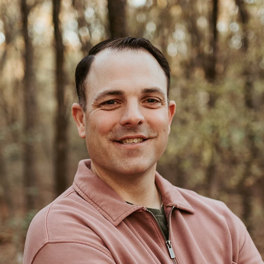 A man with short dark hair and light skin, smiling with his arms crossed, standing outdoors in a wooded area with trees and blurred background.