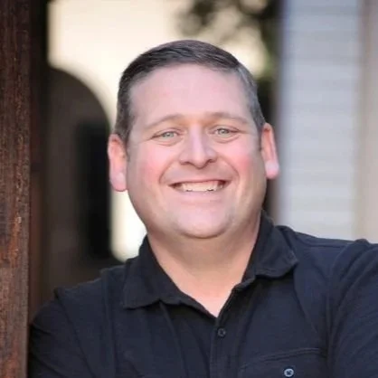 A man with short dark hair smiling against an outdoor background.