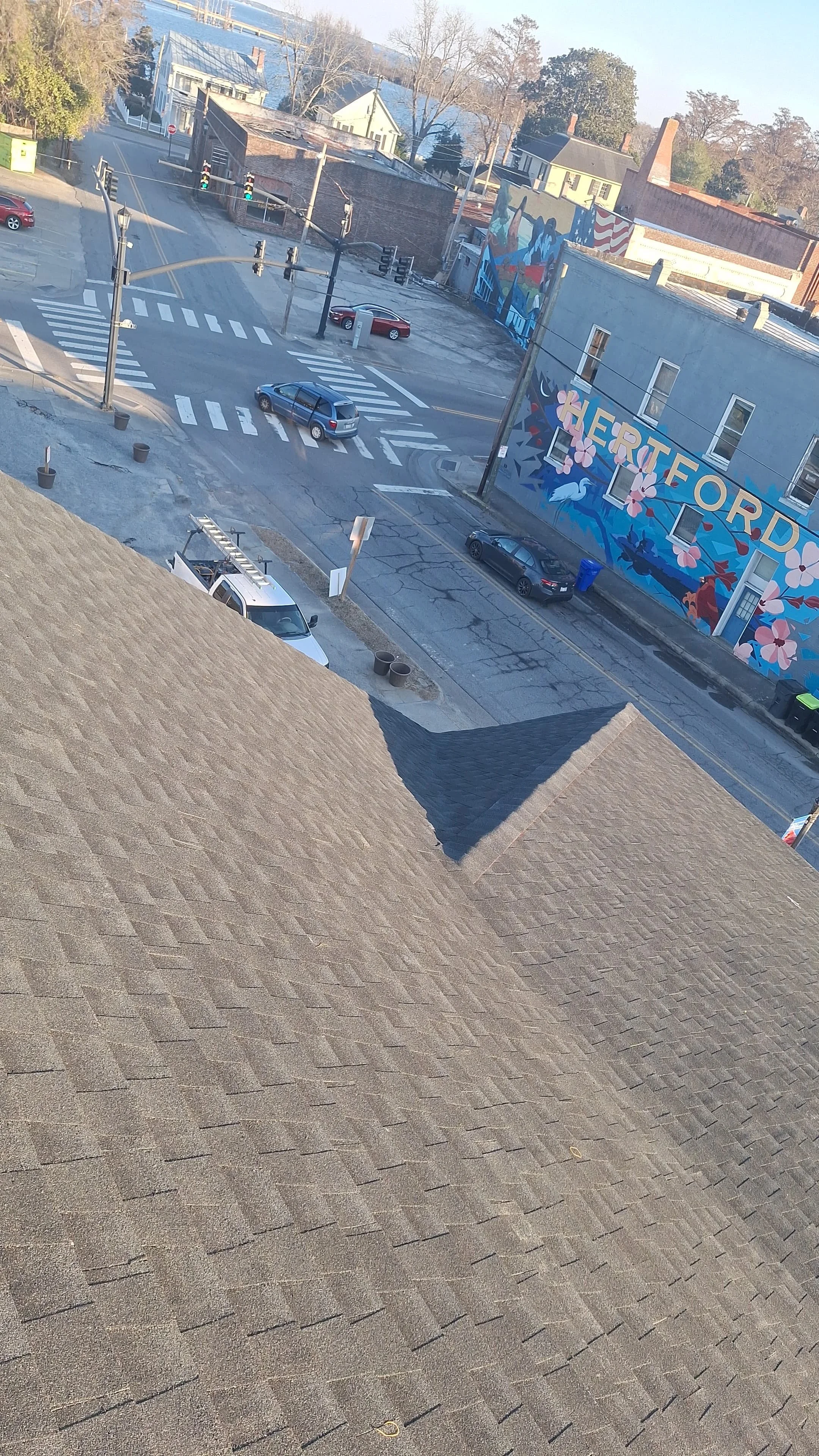 View from a rooftop shows a street intersection with several parked and moving cars, utility poles with wires, a colorful mural on the side of a building reading 'HERITORD' with floral and mountain designs, and residential buildings with trees in the background.
