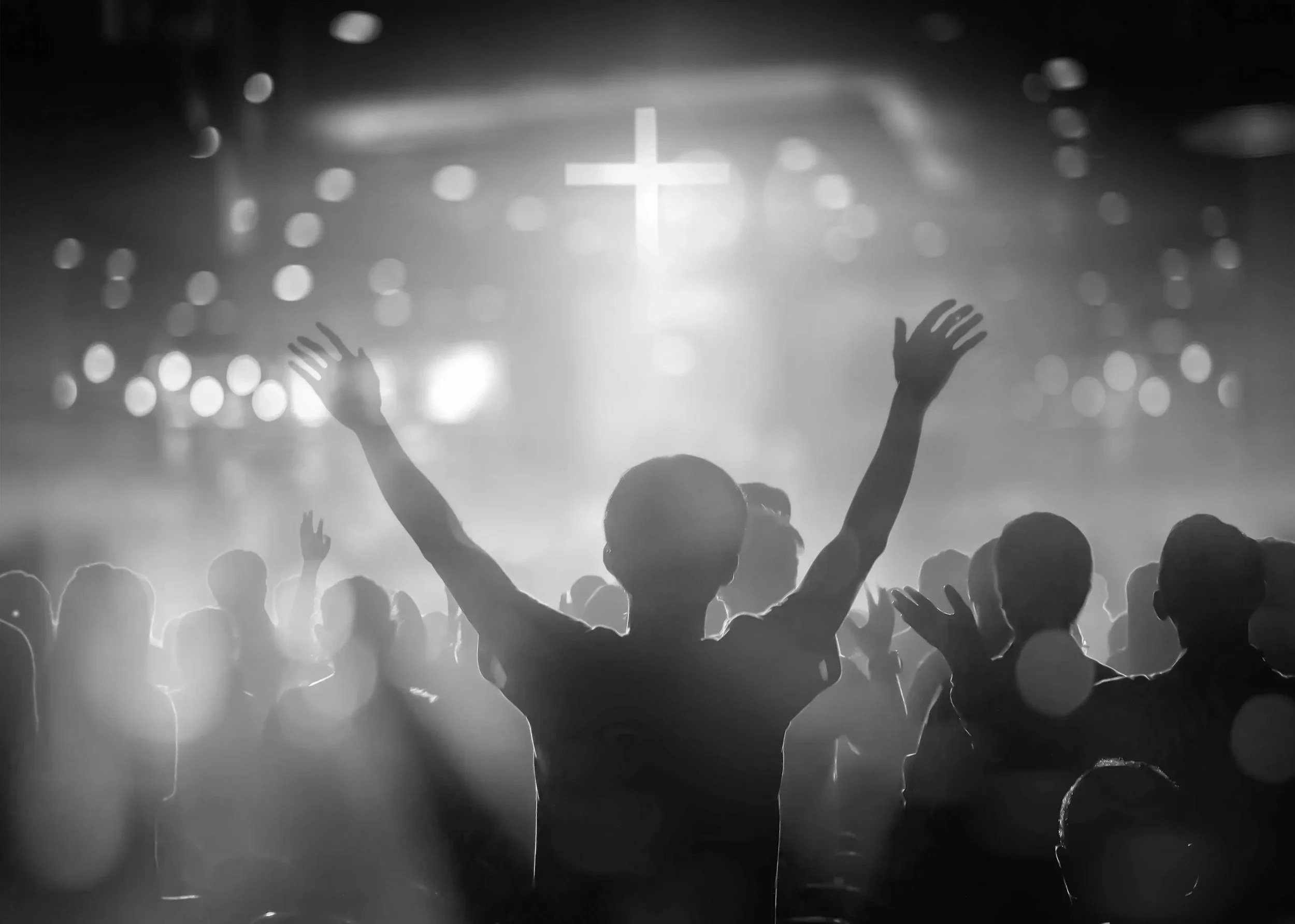 A person with arms raised in front of a crowd in a church or religious gathering, with a cross projected in the background.