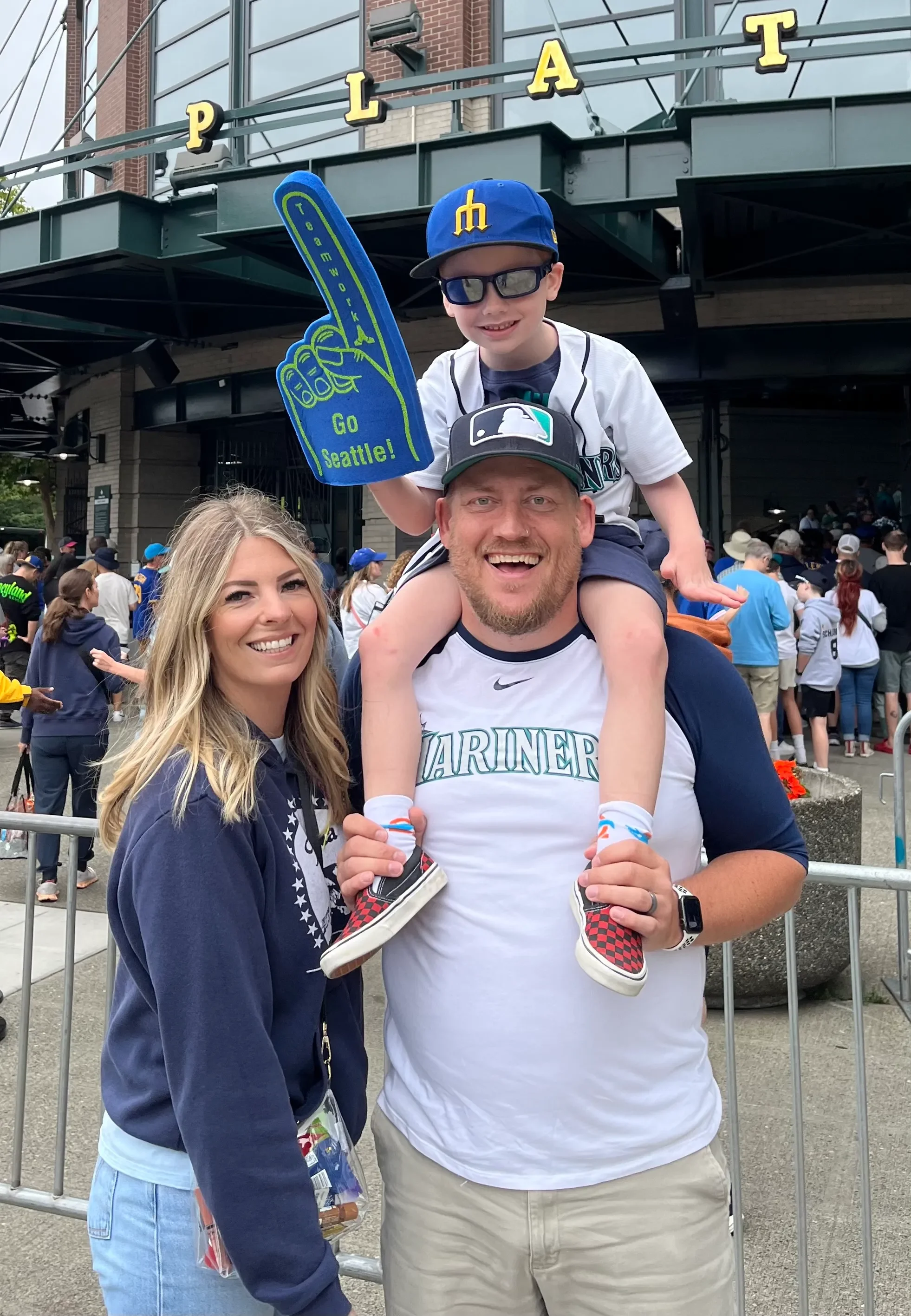 A family at a baseball game, with the man wearing a Mariners shirt, the boy on his shoulders holding a foam finger that says 'Go Seattle!' and the family standing in front of a stadium entrance with the word 'PLAT' visible.