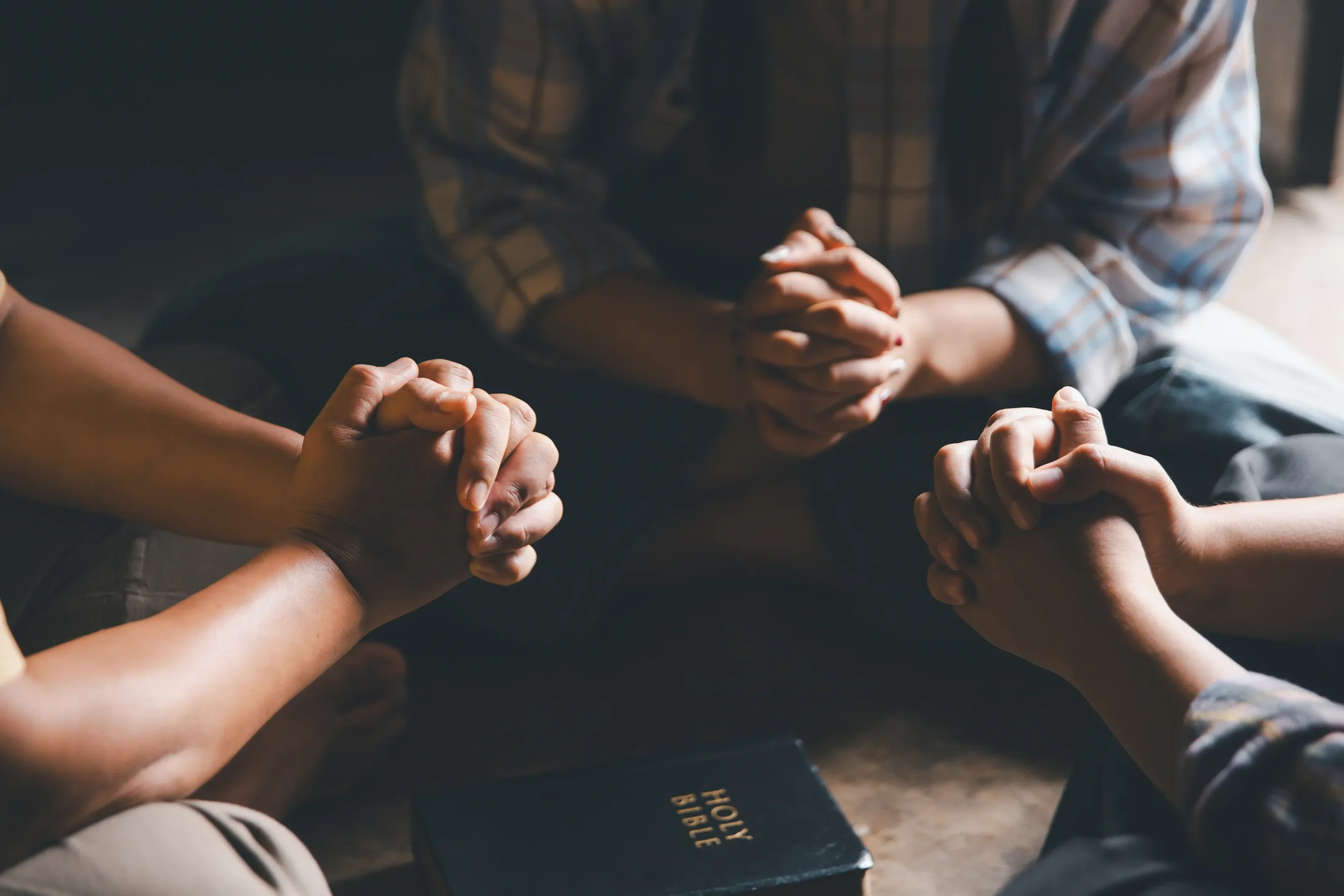 People praying with hands clasped together, seated on the floor around a Bible.