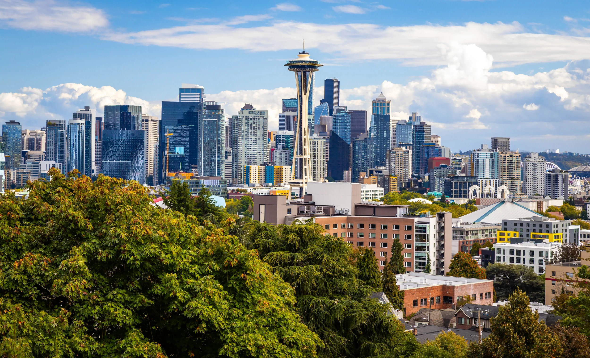 A cityscape of downtown Seattle with the Space Needle in the center, surrounded by tall skyscrapers and lush green trees in the foreground, under a partly cloudy sky.