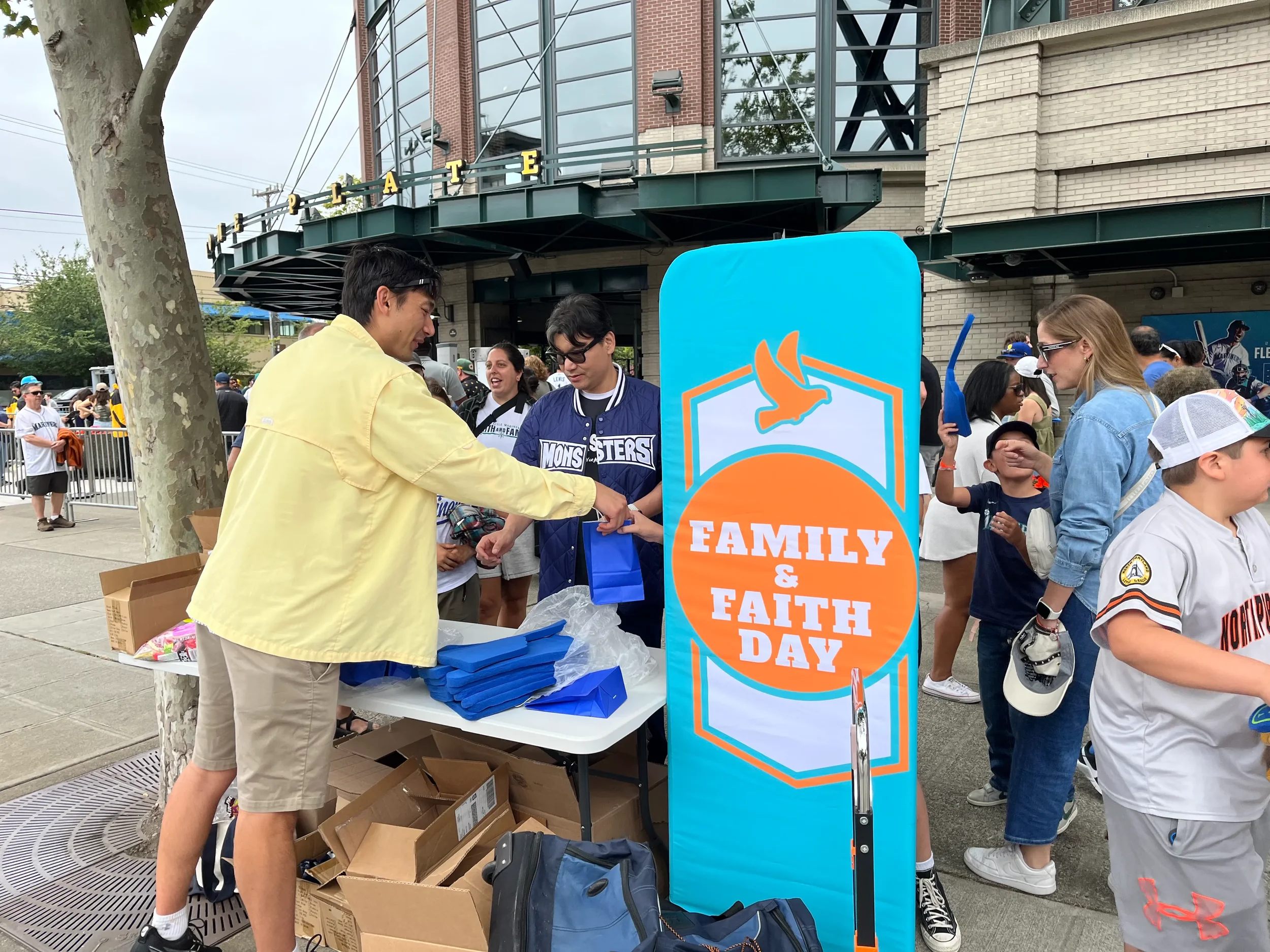 People gathered outside for Family & Faith Day event, distributing items at a booth with a large blue sign, some with children and adults, some wearing sports jerseys.