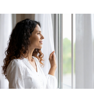 Woman with curly hair looking out a window with white curtains