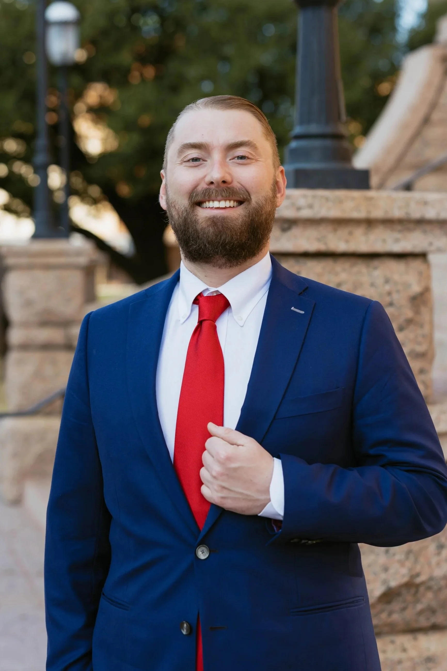Jacob Gamble, criminal defense attorney, in a blue suit, white shirt, and red tie standing outdoors in front of a stone structure and trees, smiling at the camera.