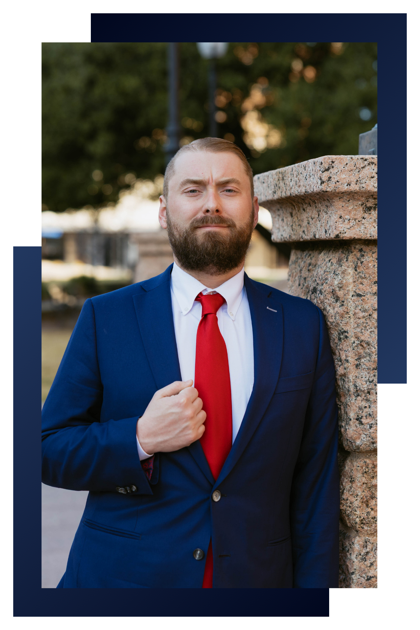 Jacob Gamble in a blue suit, white shirt, and red tie stands outdoors next to a stone structure, with trees and a park-like setting in the background.