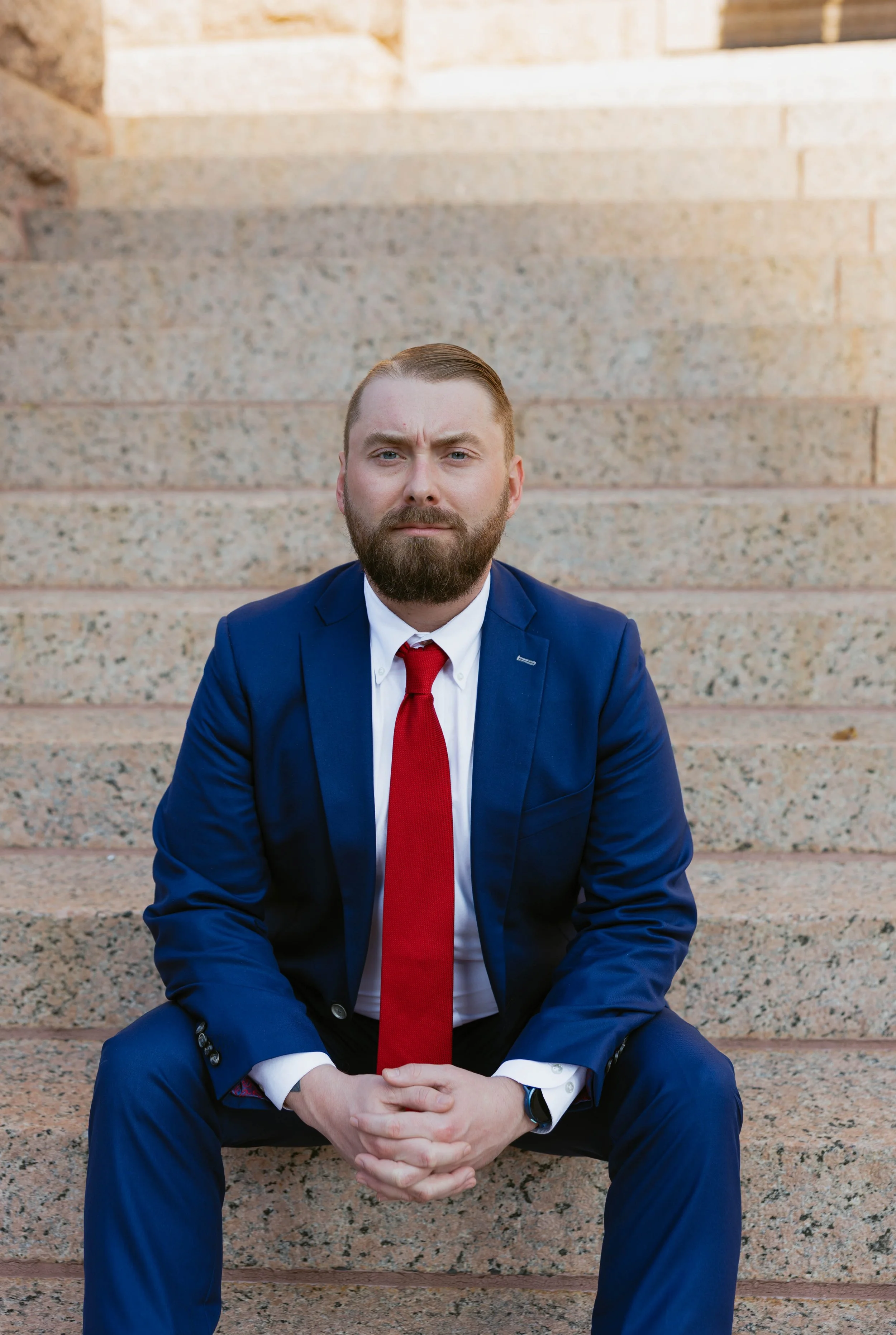 Jacob Gamble, criminal defense attorney, in blue suit and red tie sitting on stone steps with serious expression