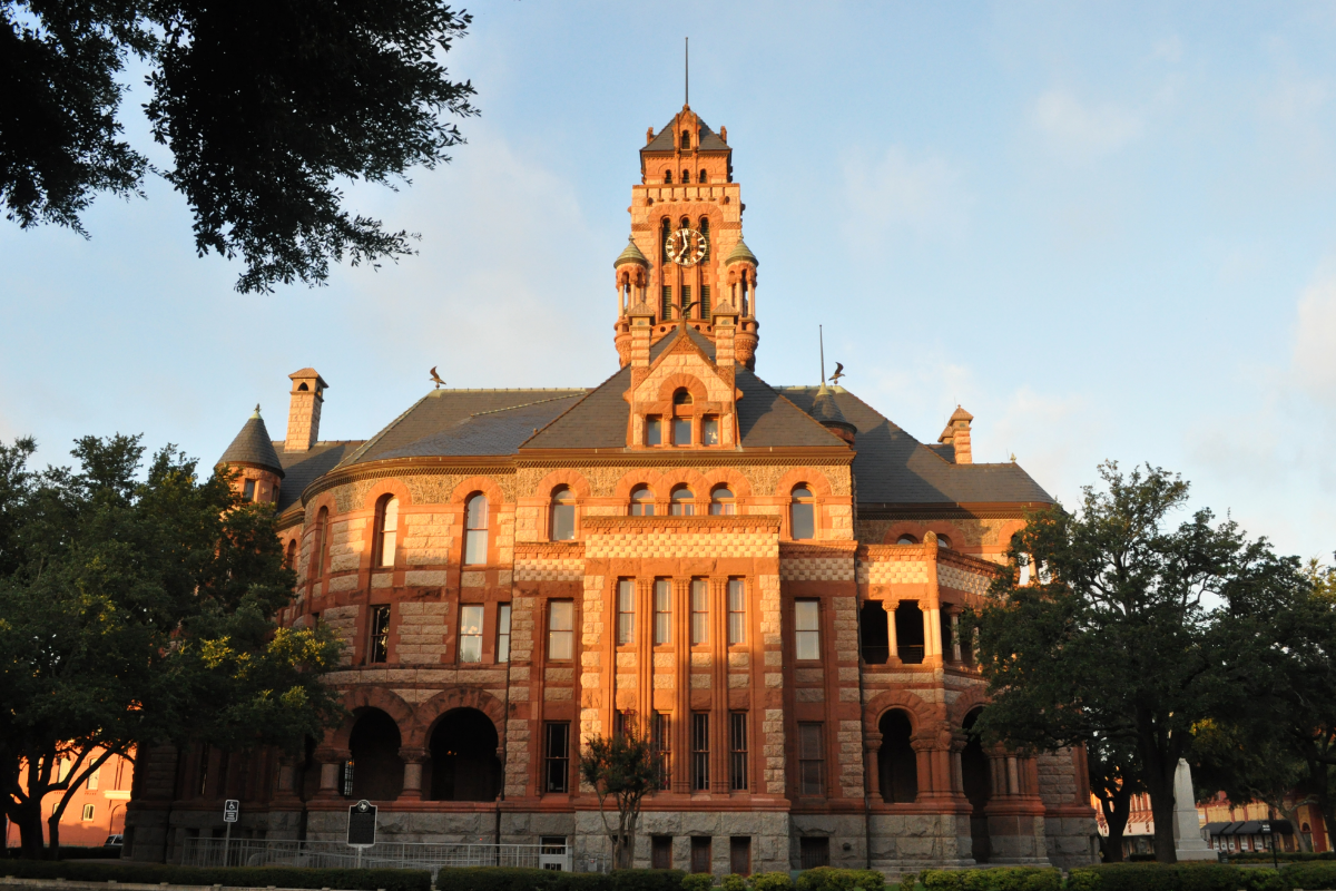 Ellis County government building with a clock tower, surrounded by trees, during the late afternoon with sunlight casting shadows.