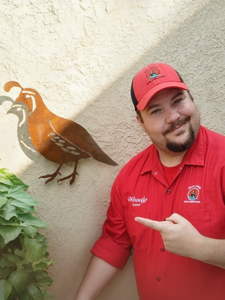 Glenn Woody Woodrome of Quail Creek Home Inspections in a red uniform with a logo and the name 'Woody' points to a wall-mounted metal bird sculpture.