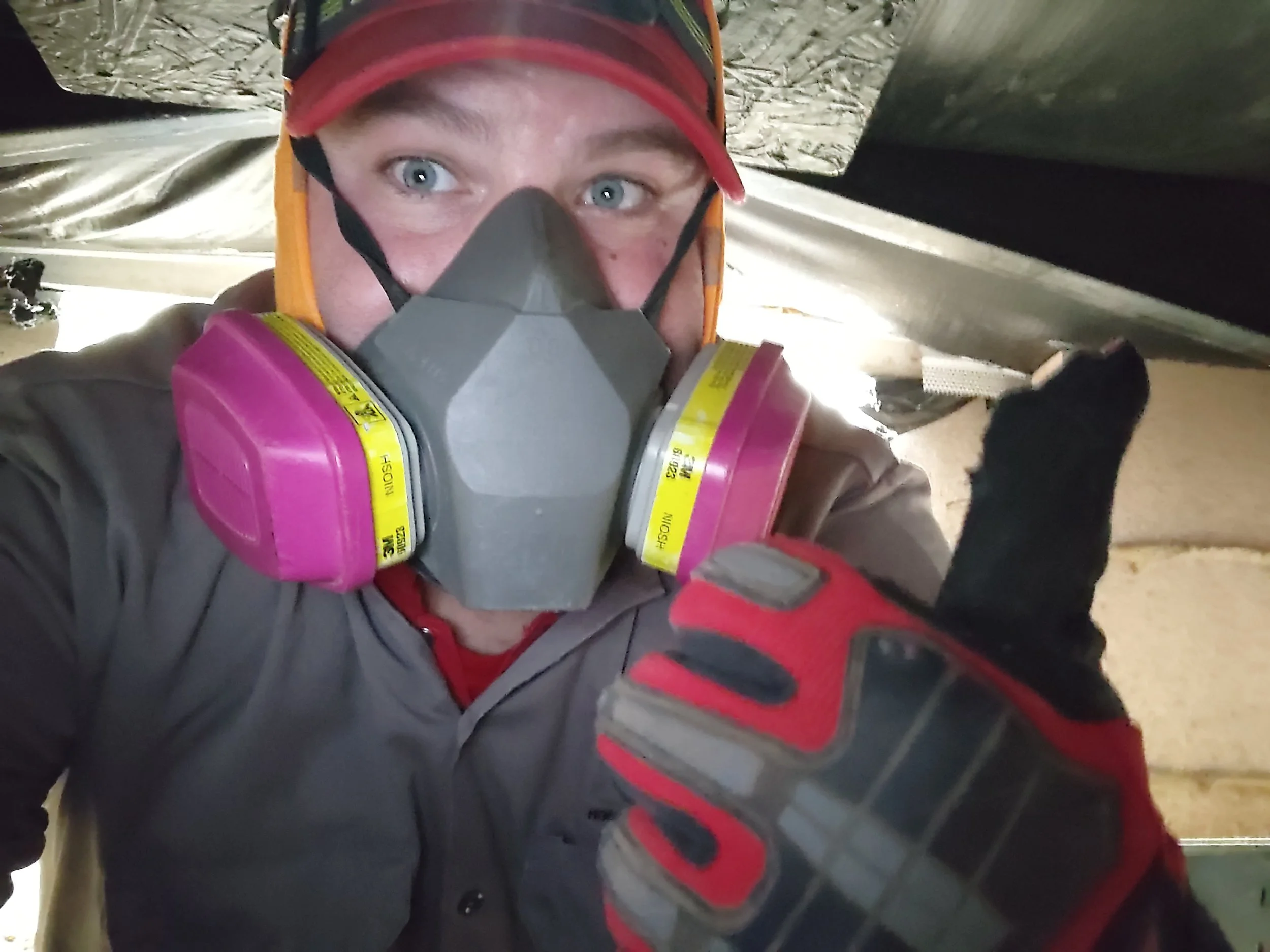 Glenn Woody Woodrome of Quail Creek Home Inspections, wearing a red hat, a gray respirator mask with pink filters, a gray jacket, and red gloves, giving a thumbs-up while in a crawlspace during a home inspection.