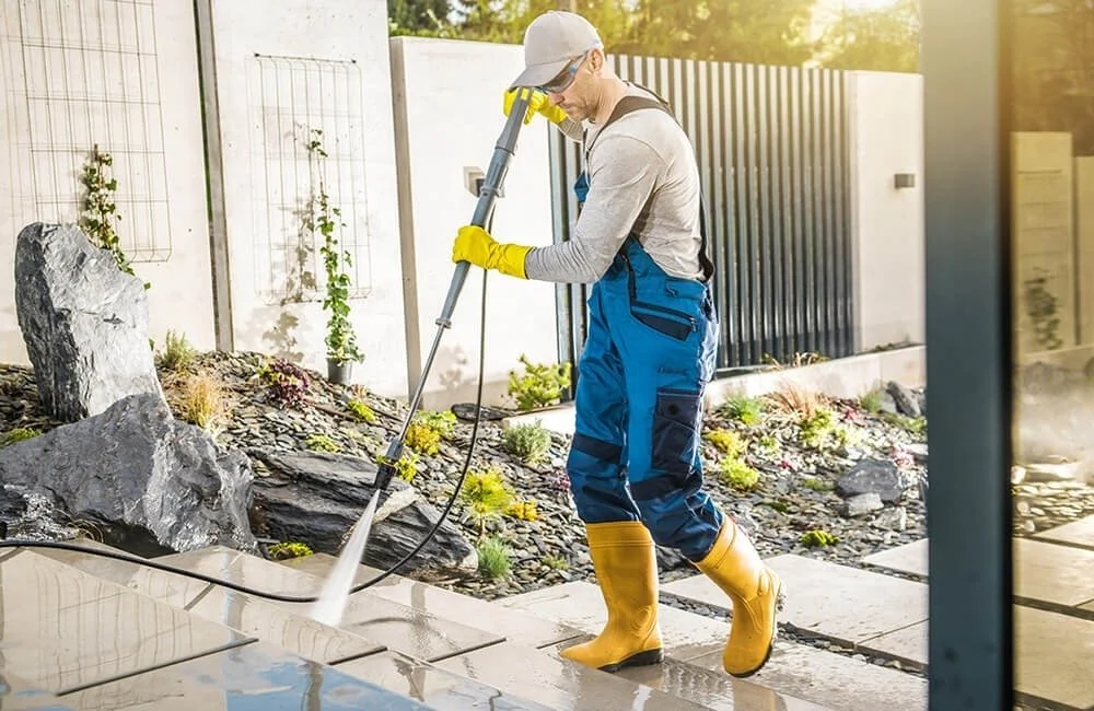 A person wearing yellow rain boots, blue overalls, and gloves power washing a concrete patio outside a house.