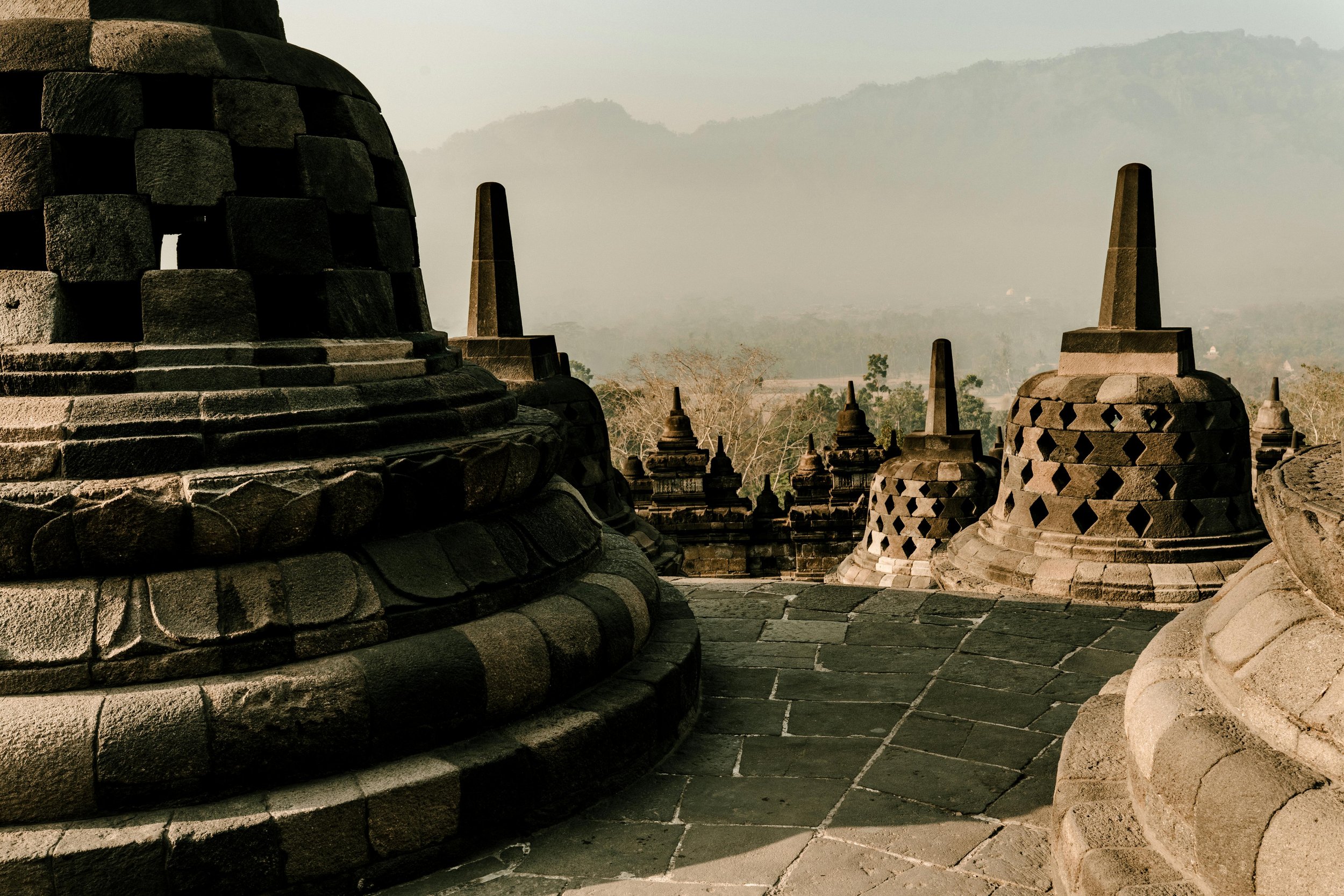 Multiple ancient stone stupas with spires on top, part of a historic temple complex, against a hazy mountain backdrop.