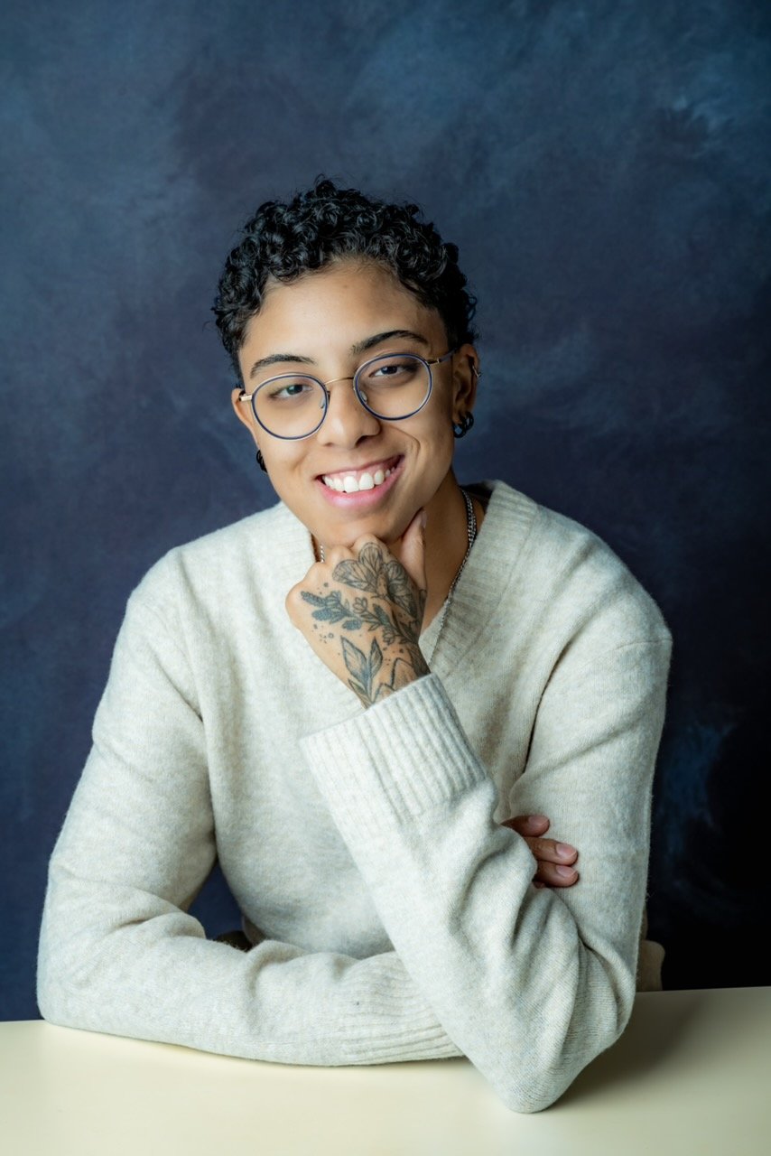A young person with short curly dark hair, glasses, and a tattooed hand resting on their chin, smiling while sitting at a table against a dark blue background.