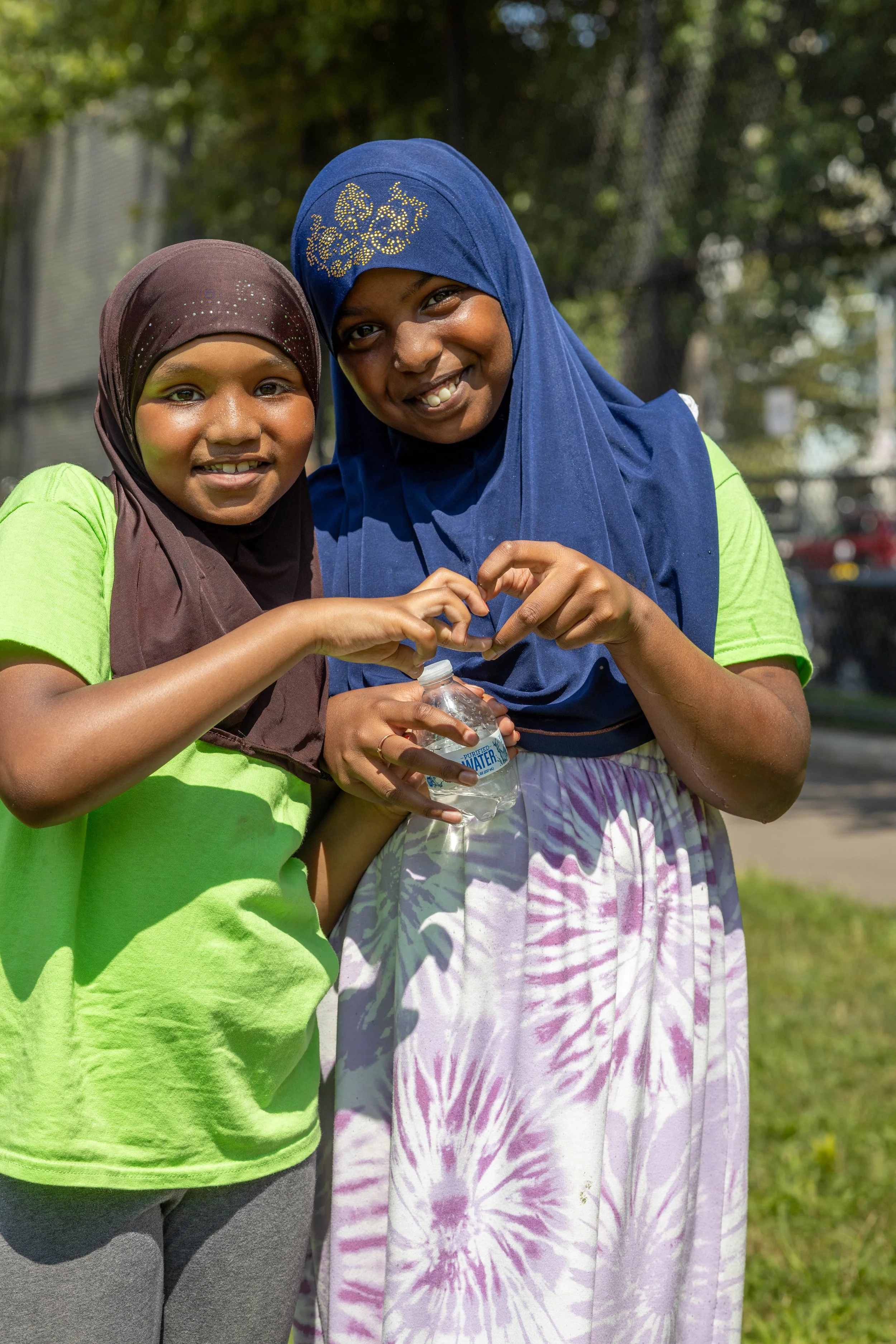 Two smiling girls wearing hijabs and bright green shirts forming a heart shape with their hands, with one holding a bottle of water, outdoors in a park.