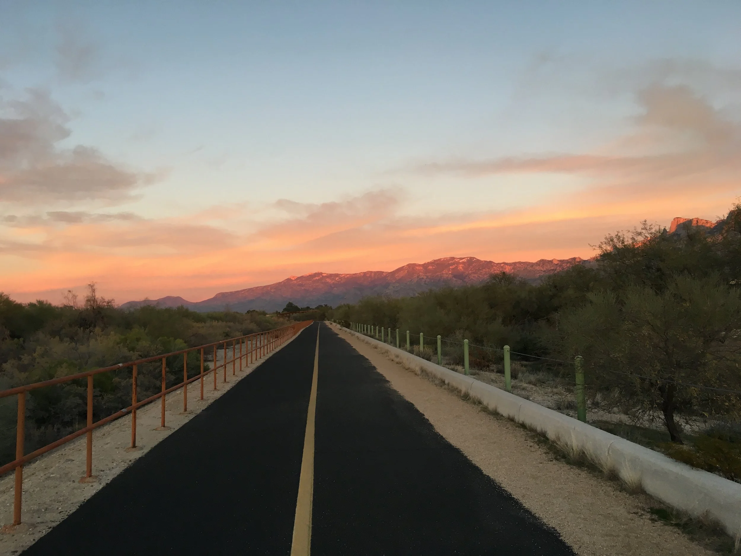 A paved road with a yellow center line extending into the distance surrounded by desert landscape with small trees and bushes, with mountains in the background at sunset or sunrise, under a partly cloudy sky.