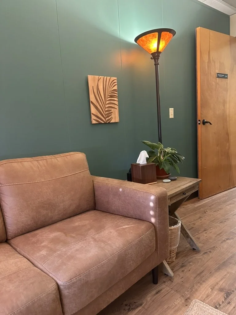 Cozy therapist's office with a brown leather couch, small wooden side table holding a plant, tissues, green wall with a leaf art piece, tall lamp with orange light, wooden door, and hardwood floor.