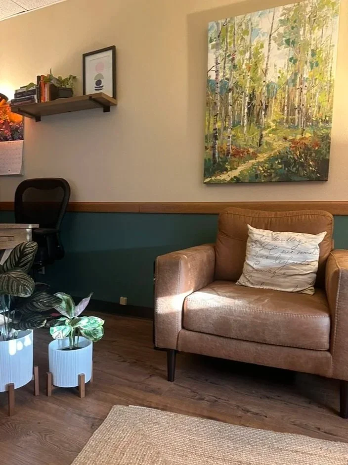A cozy therapist's office with a tan leather armchair that has a white decorative pillow, wooden flooring, potted plants on the floor, and a large colorful painting of a forest on the wall.