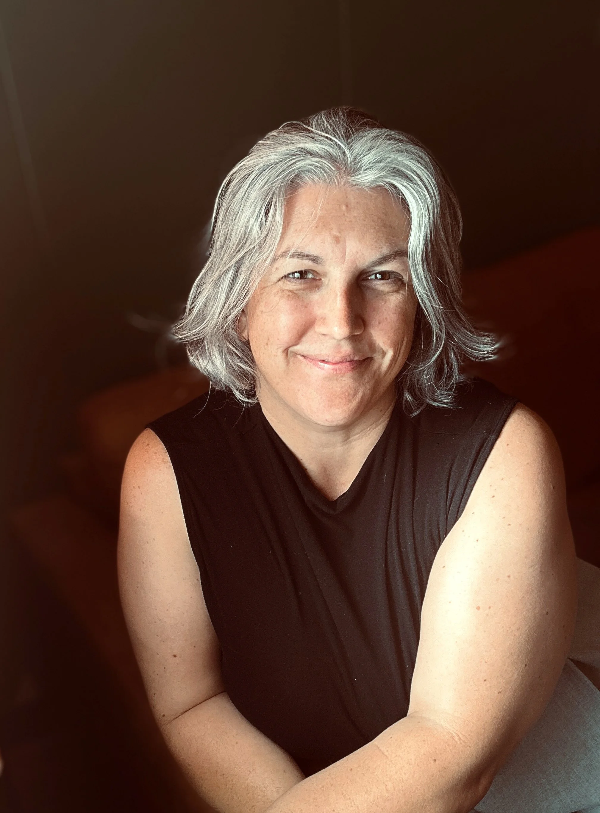 Close-up of a woman with short, gray and white hair, smiling, wearing a black sleeveless top, with fair skin and freckles, sitting with arms crossed, soft lighting, dark background.