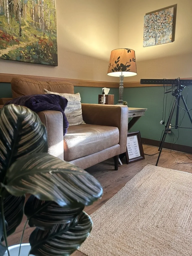 A cozy therapy space with a tan leather sofa, a wooden side table with a lamp, artwork on the walls, a framed quote, a plant in the foreground, and a beige rug on wooden flooring.